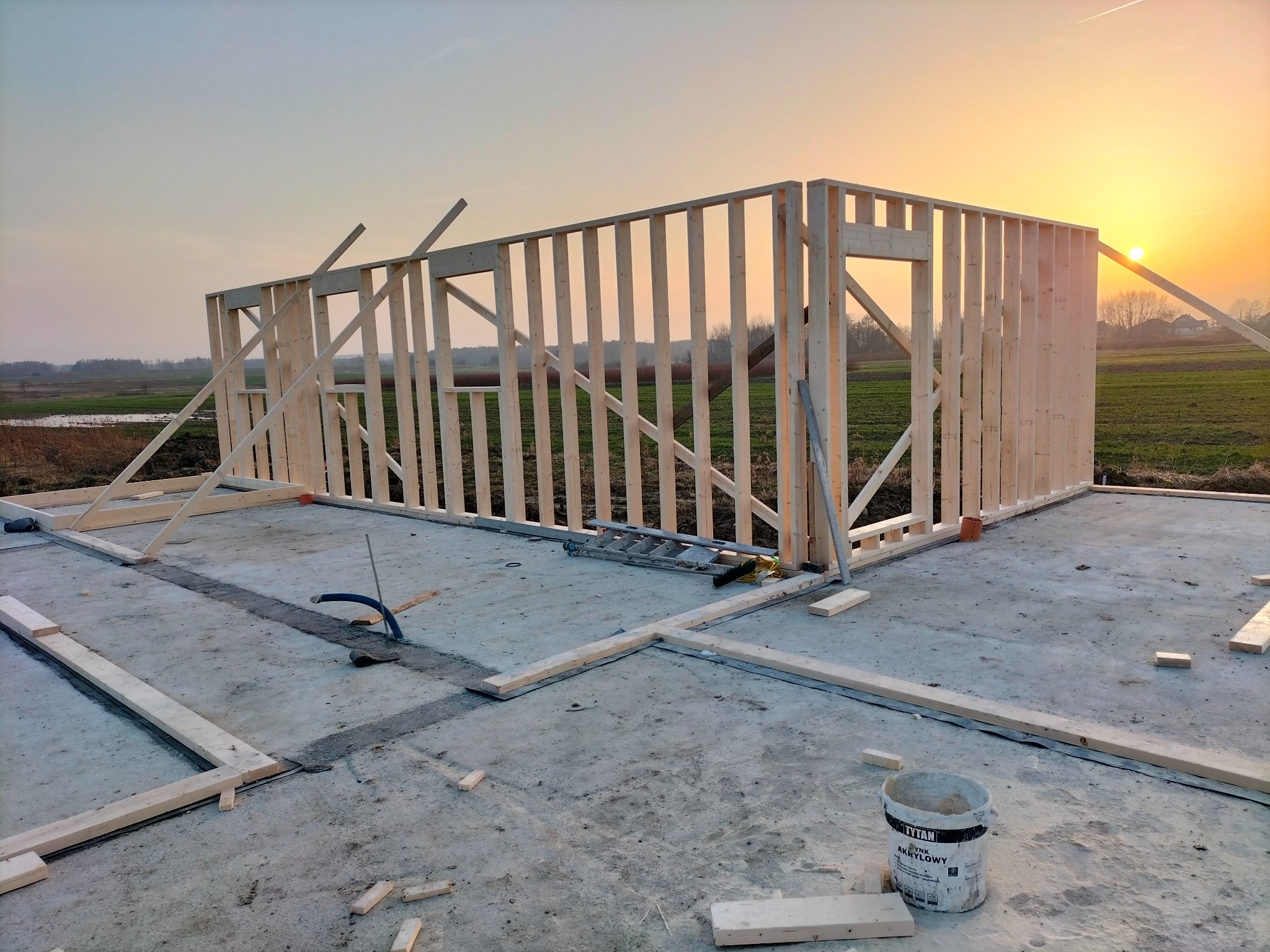 Wczesna faza budowy domu szkieletowego: drewniana konstrukcja ścian na fundamencie, widok na zachodzące słońce i rozległe pole. Na fundamencie leżą deski i stoi drabina.