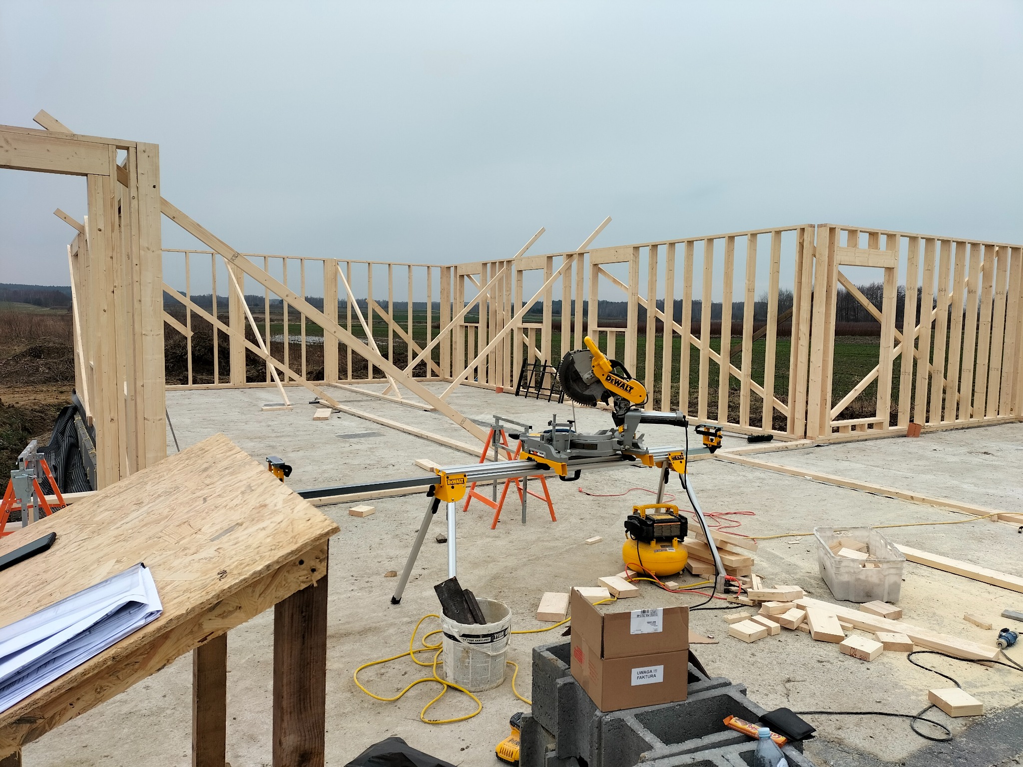 Wznoszenie konstrukcji szkieletowej domu na fundamencie. Widoczna piła DeWalt, kompresor i dokumentacja. Ściany z drewna w trakcie montażu, krajobraz w tle.