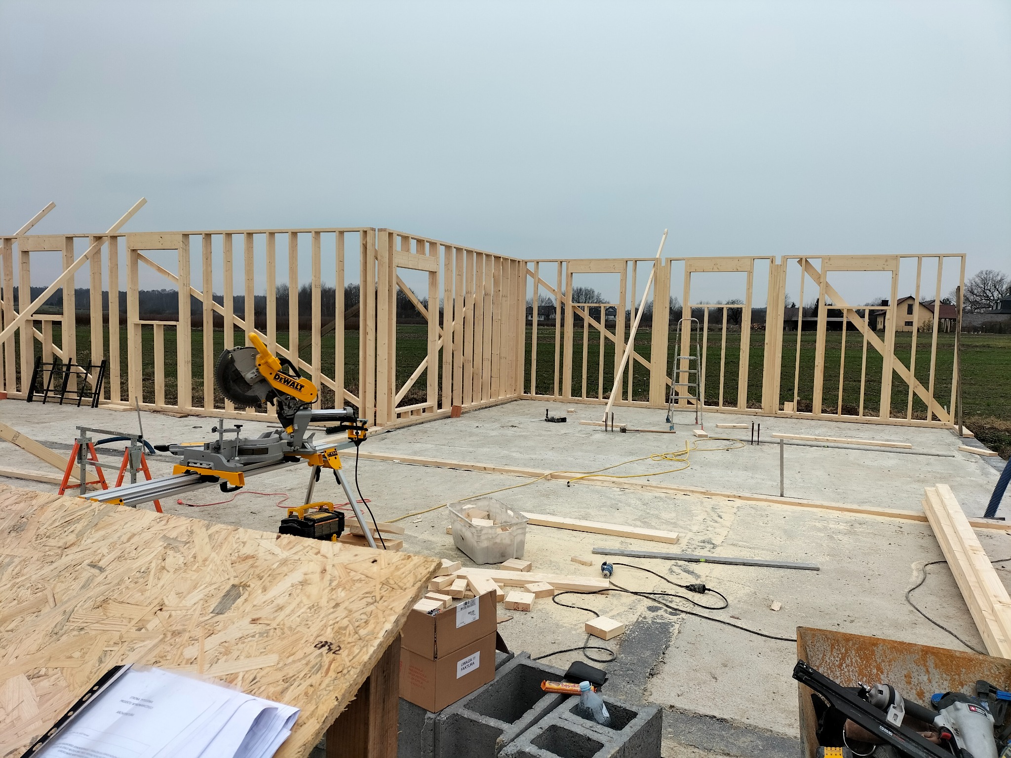 Wznoszenie szkieletu domu z drewna na betonowej płycie fundamentowej. Widoczne narzędzia: piła ukośnica DeWalt, poziomica i elementy konstrukcyjne ścian. Etap budowy.