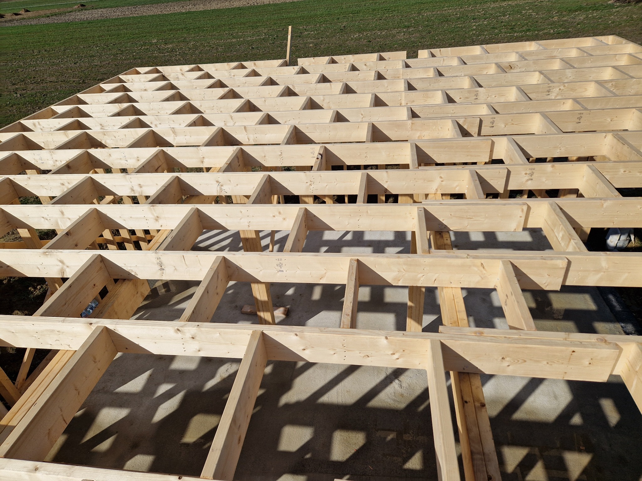 Konstrukcja podłogi szkieletowej w trakcie budowy, widok z góry, jasne drewno, cień belek na podłożu, trawa w tle. Elementy złączone gwoździami.