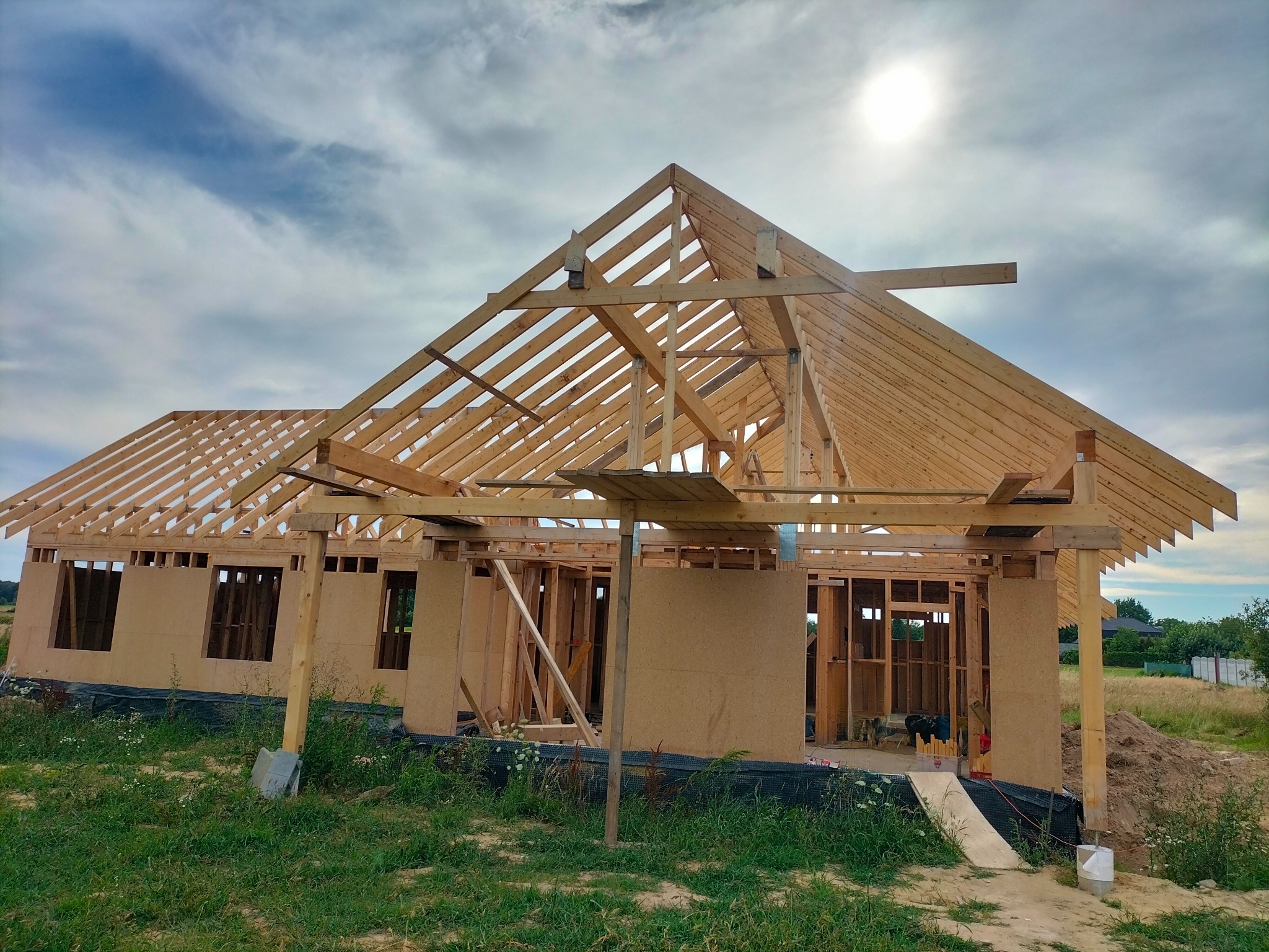 Budowa domu szkieletowego w trakcie realizacji: widoczne drewniane elementy konstrukcyjne ścian i dachu na tle błękitnego nieba z chmurami.
