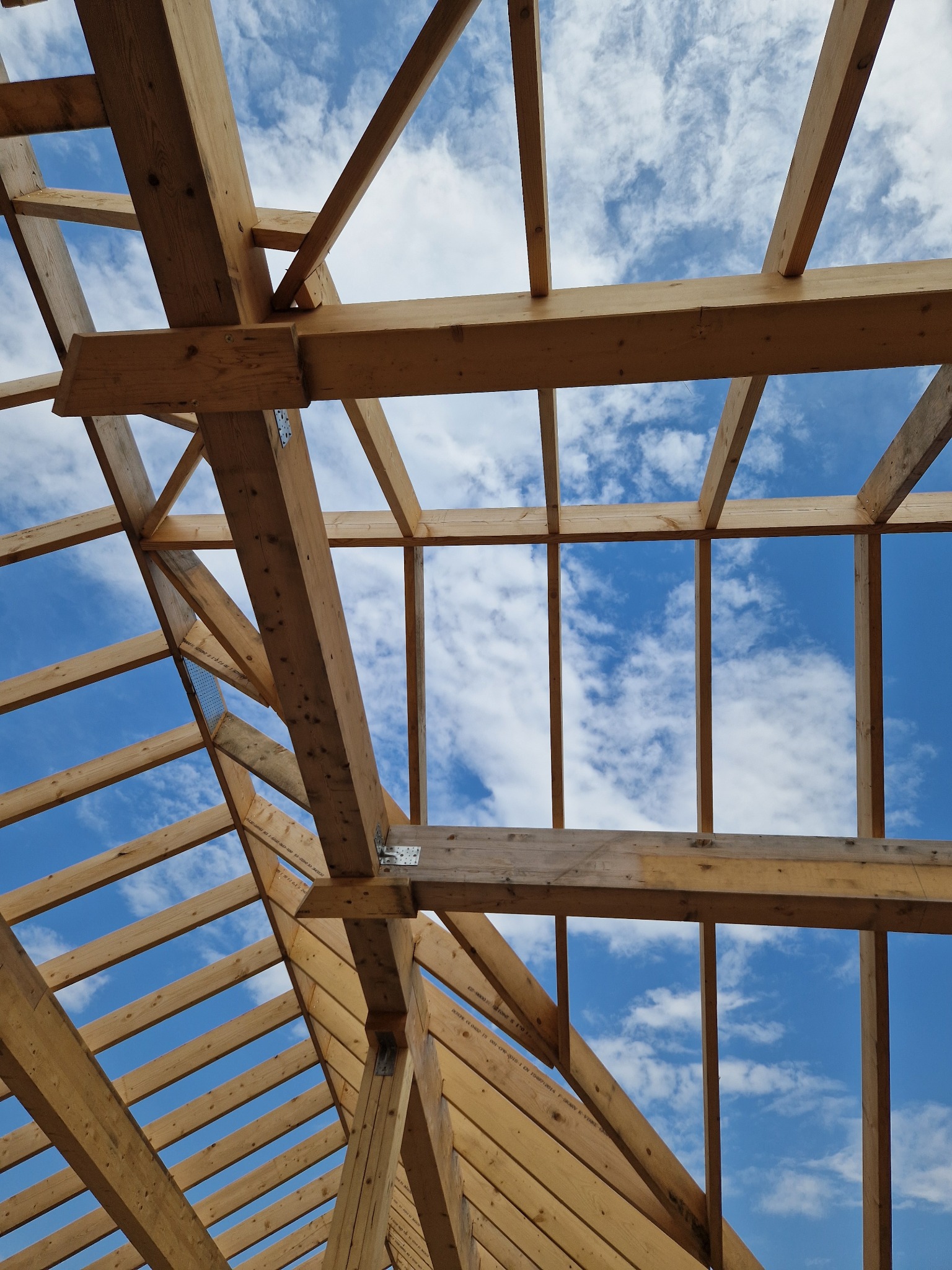 Konstrukcja drewniana dachu z widocznymi elementami łączącymi na tle błękitnego nieba z chmurami. Drewno w jasnym odcieniu. Kąt patrzenia z dołu, w górę.