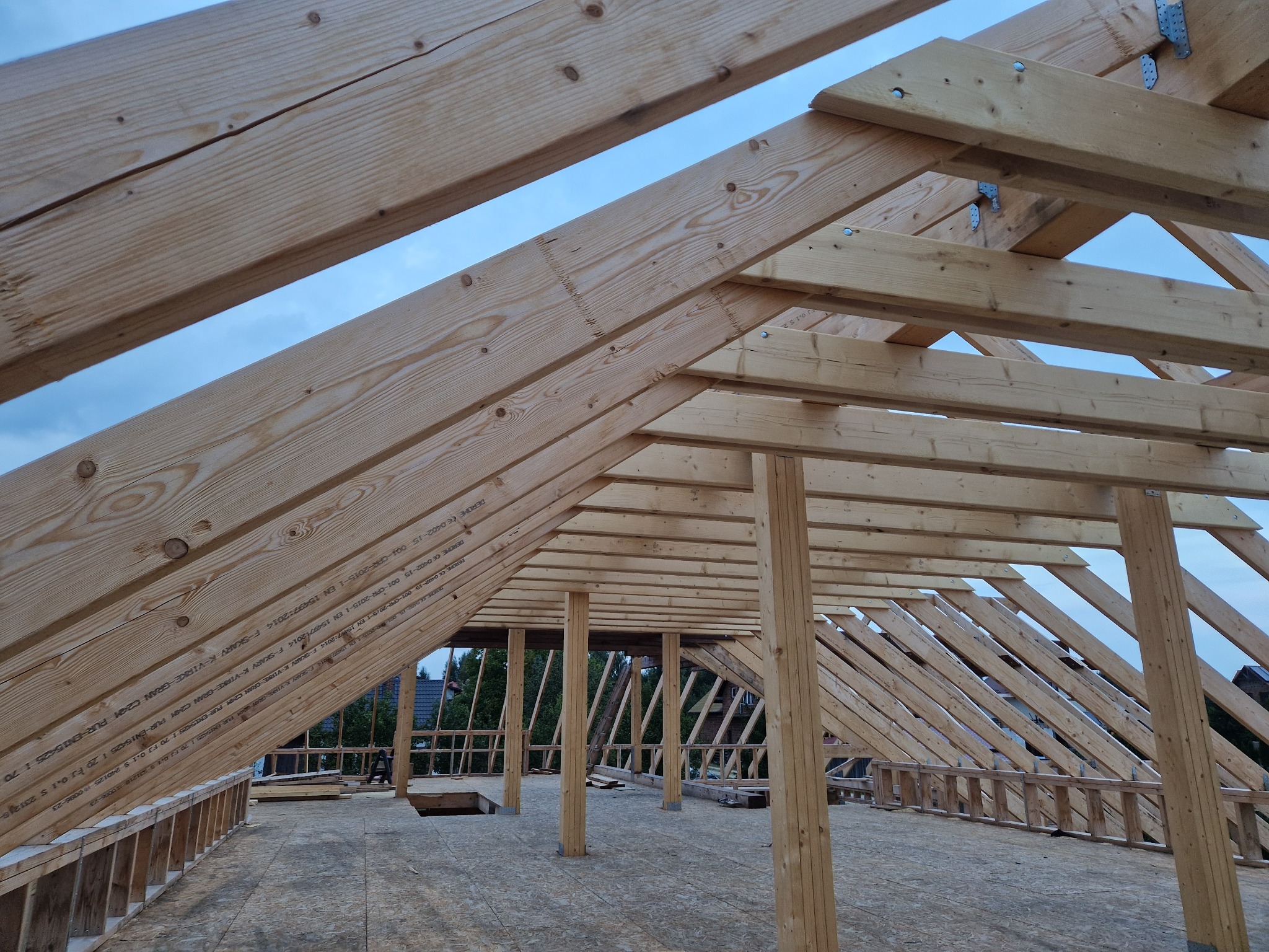 Konstrukcja dachu z drewnianych belek, widok z wnętrza budynku w trakcie budowy. Widoczne elementy złączne i oznaczenia na drewnie.
