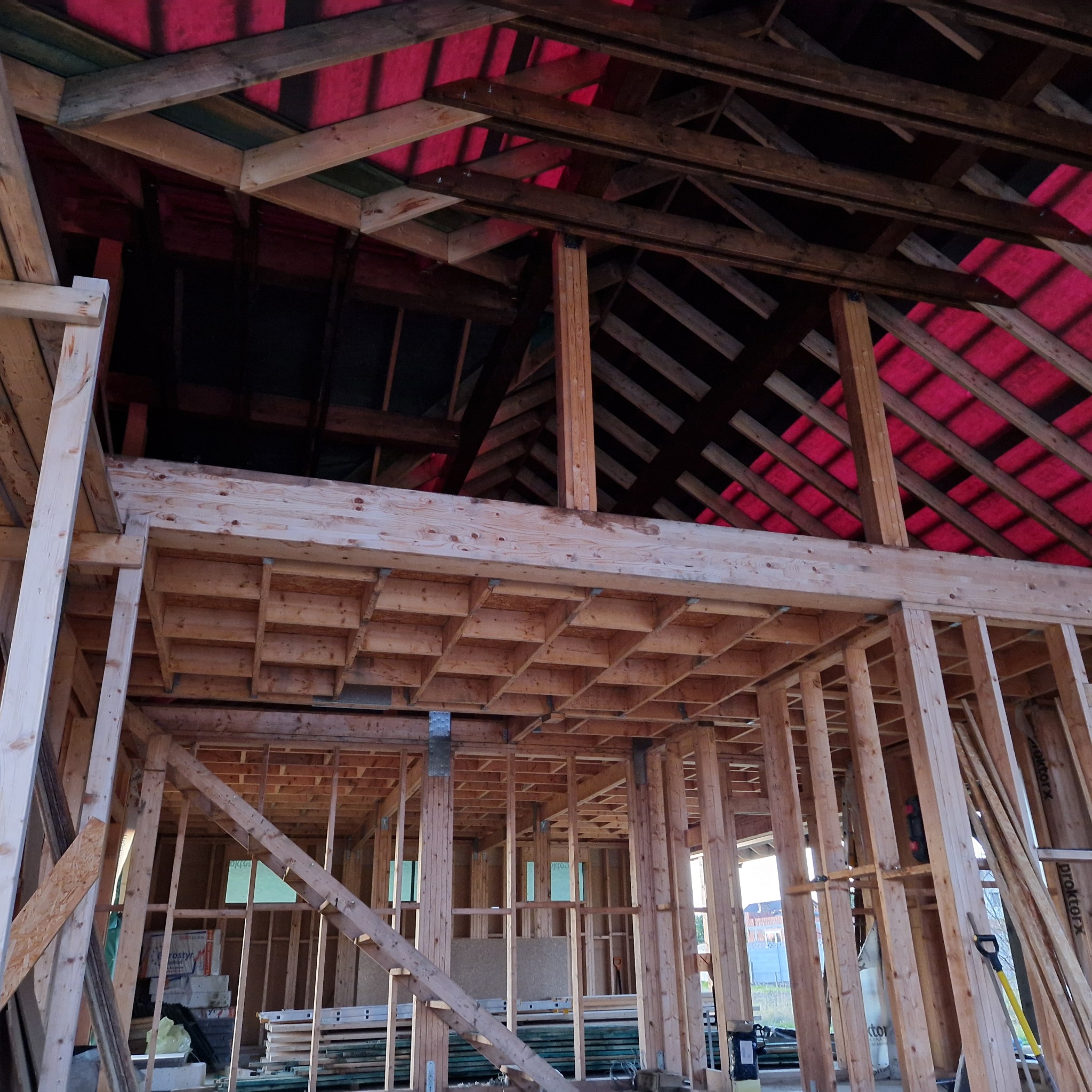 Wnętrze domu szkieletowego w trakcie budowy. Widoczne drewniane elementy konstrukcyjne, czerwona membrana dachowa i częściowo ułożone podłogi. Surowy stan.