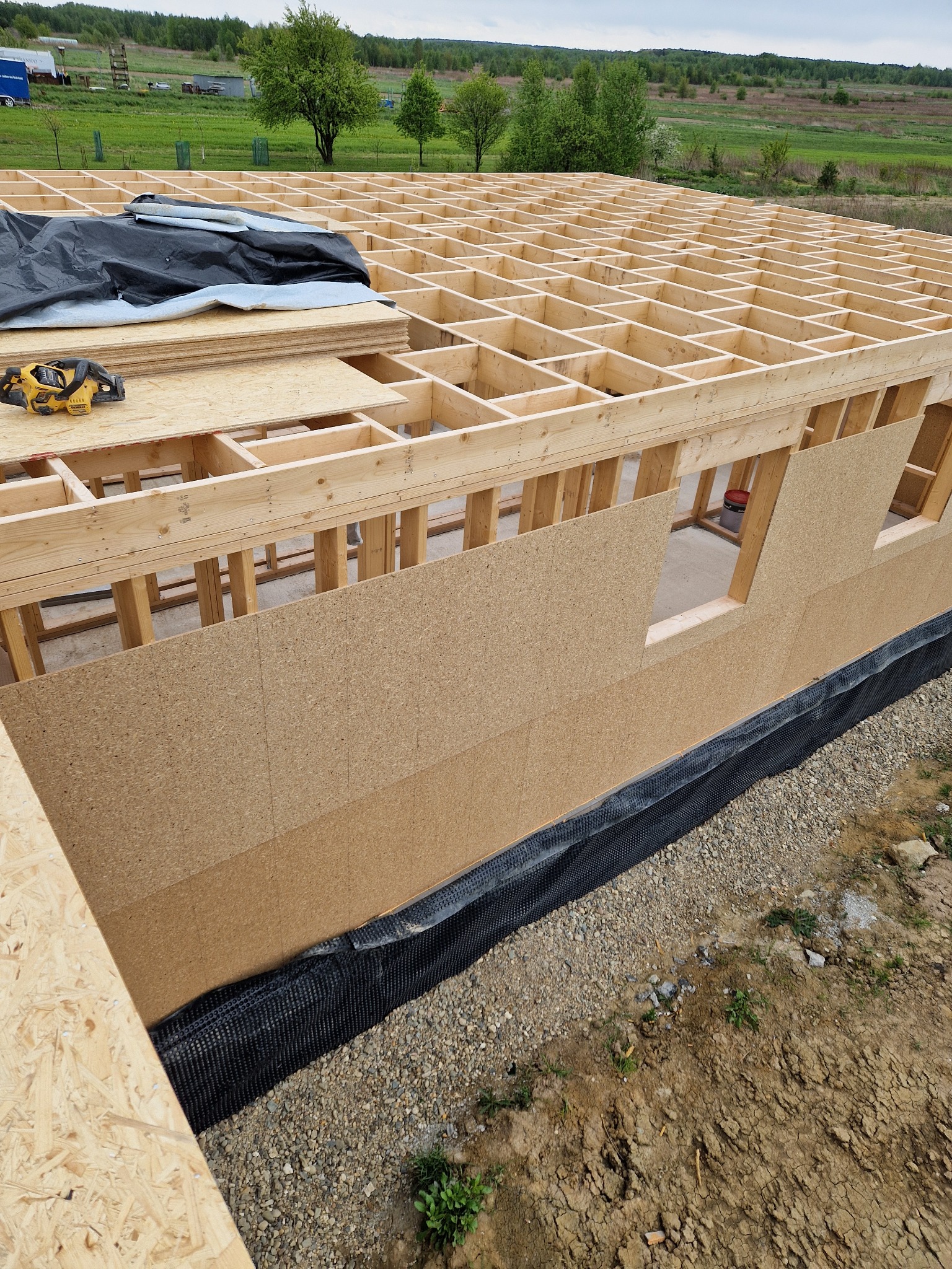 Wznoszenie domu: szkielet drewniany z płytami OSB, widoczne narzędzia i izolacja. Budowa na tle zieleni, widać strukturę dachu i otwory okienne w ścianach.