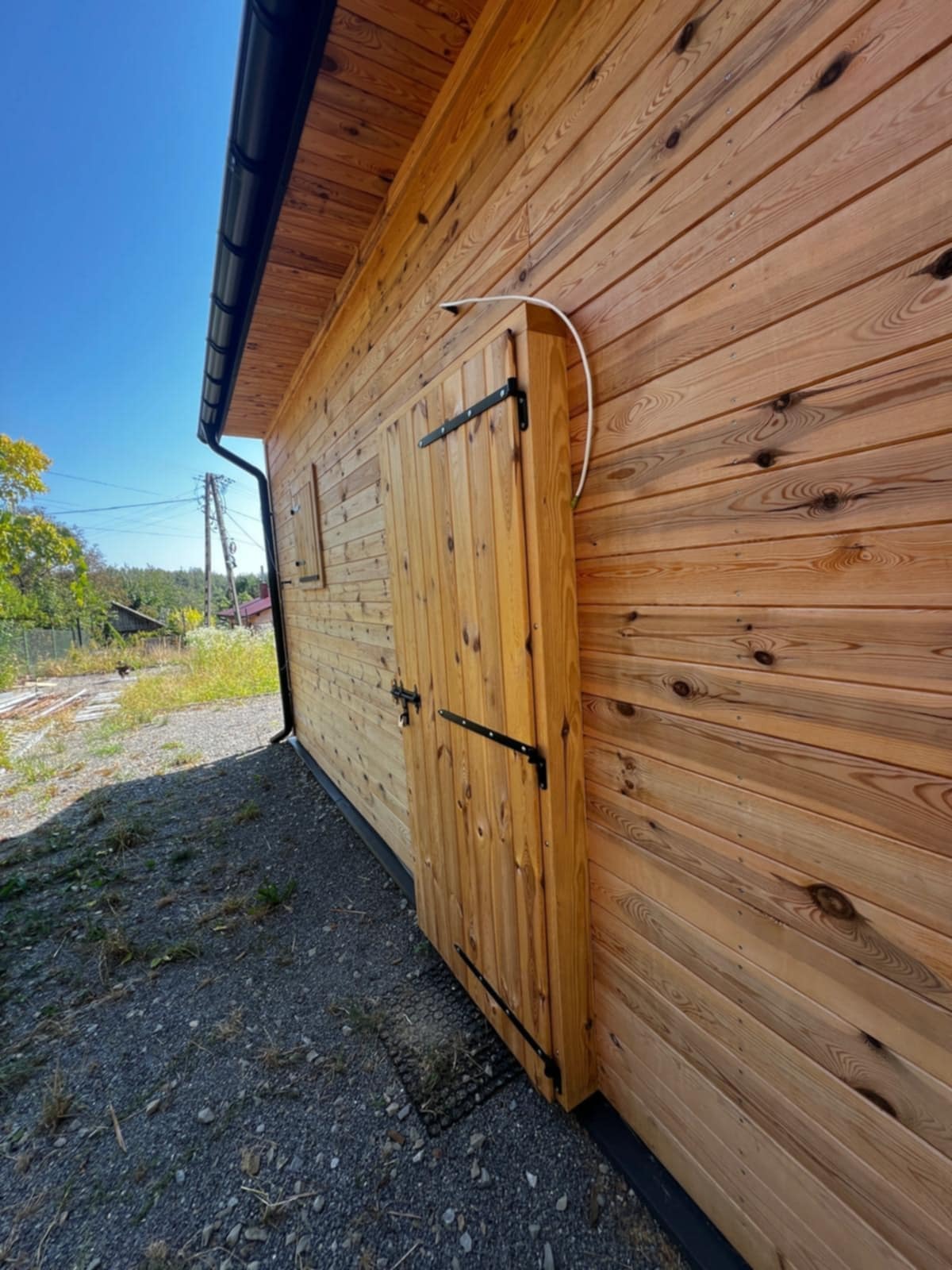 Ściana budynku z drewna z drzwiami i małym oknem, widok z zewnątrz, z widocznym fragmentem dachu i otoczenia. Charakterystyczne czarne okucia drzwi.