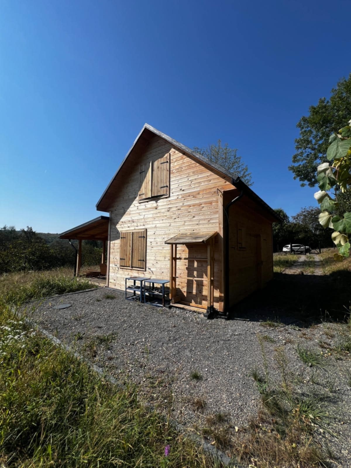 Jasny, drewniany dom szkieletowy z okiennicami, otoczony zielenią i żwirowym podłożem w słoneczny dzień. Widoczny fragment zadaszonego tarasu i dodatkowe wejście.