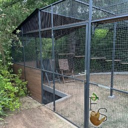 Folding System Dominik Sajdak - Woliera z tunelem i półkami, wykonana z metalowej siatki. Część wolierowa zadaszona, wewnątrz krzesła. Widoczne logo firmy na ogrodzeniu.