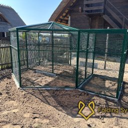 Folding System Dominik Sajdak - Sześciokątna, zielona woliera z siatki na ziemi, z widocznymi elementami konstrukcyjnymi i tłem wiejskiego krajobrazu. Widoczny znak wodny.