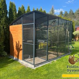 Folding System Dominik Sajdak - Zewnętrzna, duża woliera z drewnianym panelem i metalową siatką, na trawniku w ogrodzie, z drzewami w tle, oświetlona słońcem.