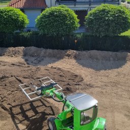 Folding System Dominik Sajdak - Zielona minikoparka Avant z osprzętem do układania kostki brukowej na przygotowanym terenie, w tle dom z czerwonym dachem i kuliste krzewy.