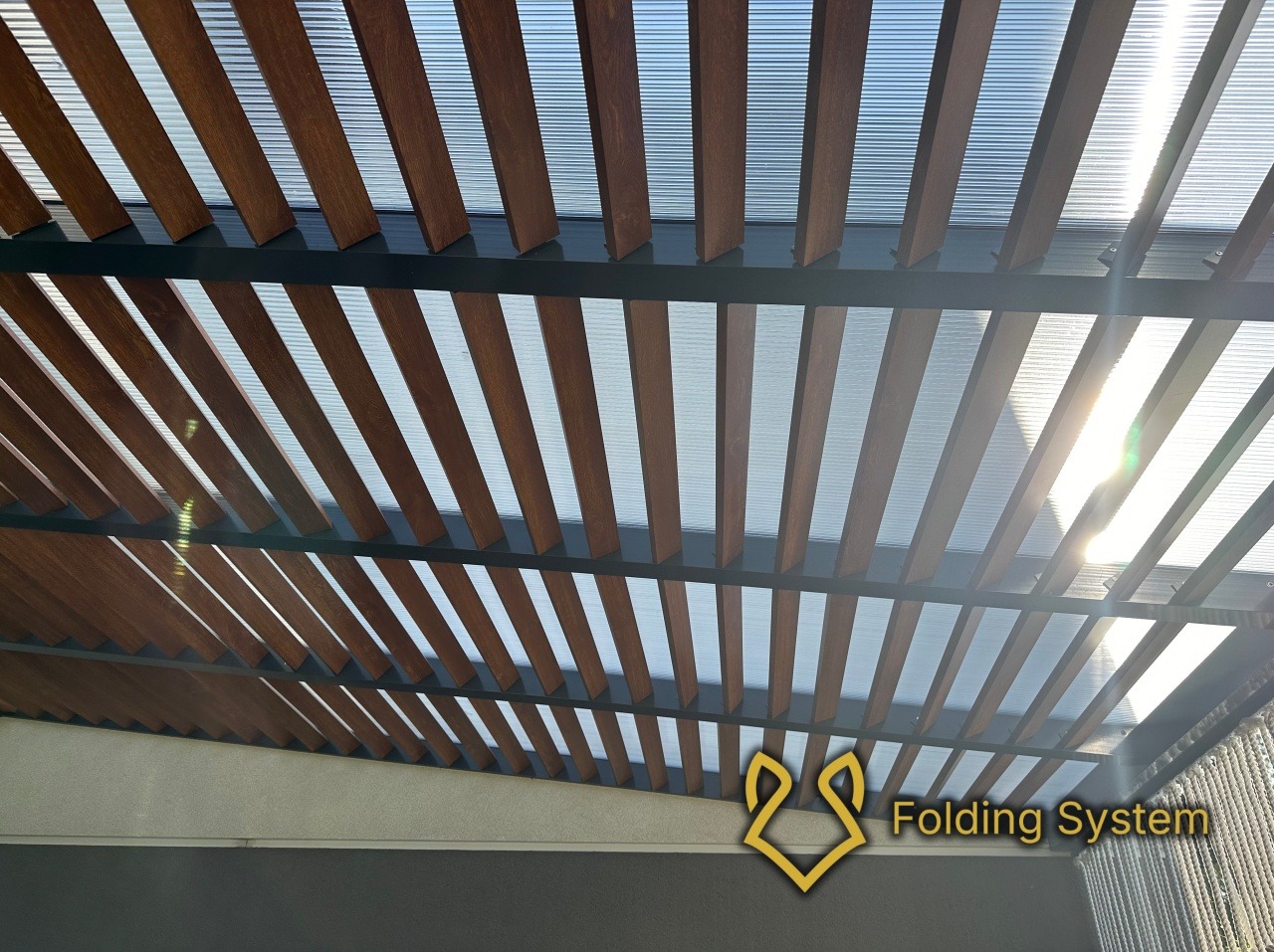 Nowoczesna pergola z drewnopodobnymi lamelami i transparentnym zadaszeniem, widziana od dołu. Słońce prześwituje przez zadaszenie, tworząc jasne refleksy. Widoczne logo firmy.