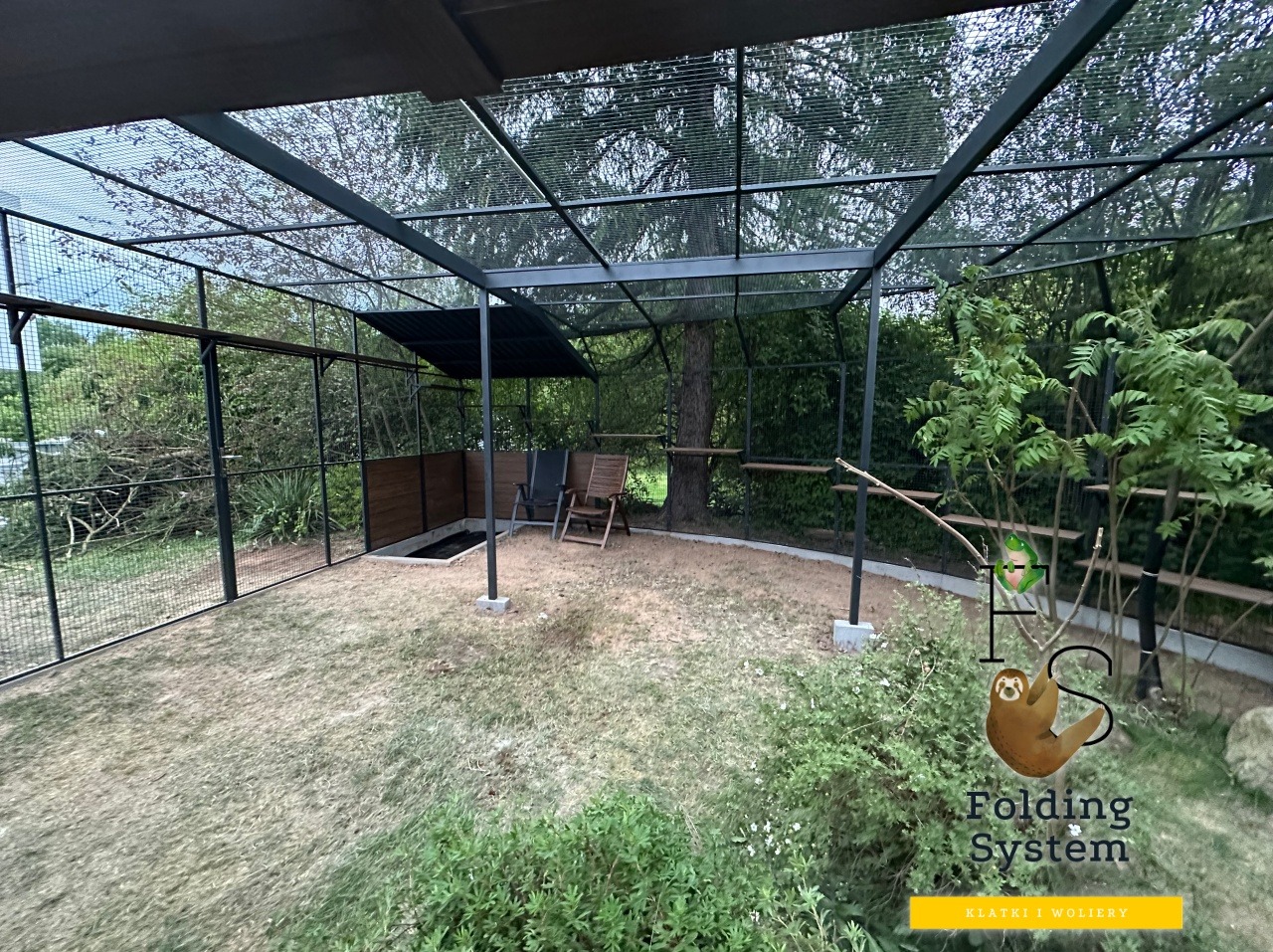 Lakierowana woliera z zadaszeniem i tunelem, idealna dla kota, otoczona zielenią. Widoczne półki i elementy konstrukcyjne. Całość wkomponowana w naturalne otoczenie.