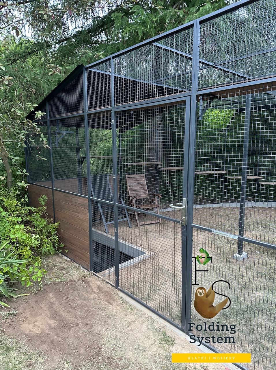 Woliera z tunelem i półkami, wykonana z metalowej siatki. Część wolierowa zadaszona, wewnątrz krzesła. Widoczne logo firmy na ogrodzeniu.