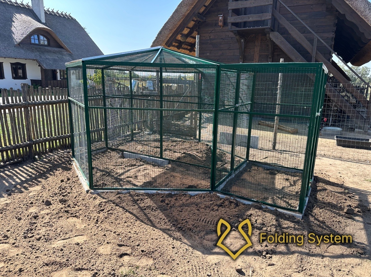 Sześciokątna, zielona woliera z siatki na ziemi, z widocznymi elementami konstrukcyjnymi i tłem wiejskiego krajobrazu. Widoczny znak wodny.