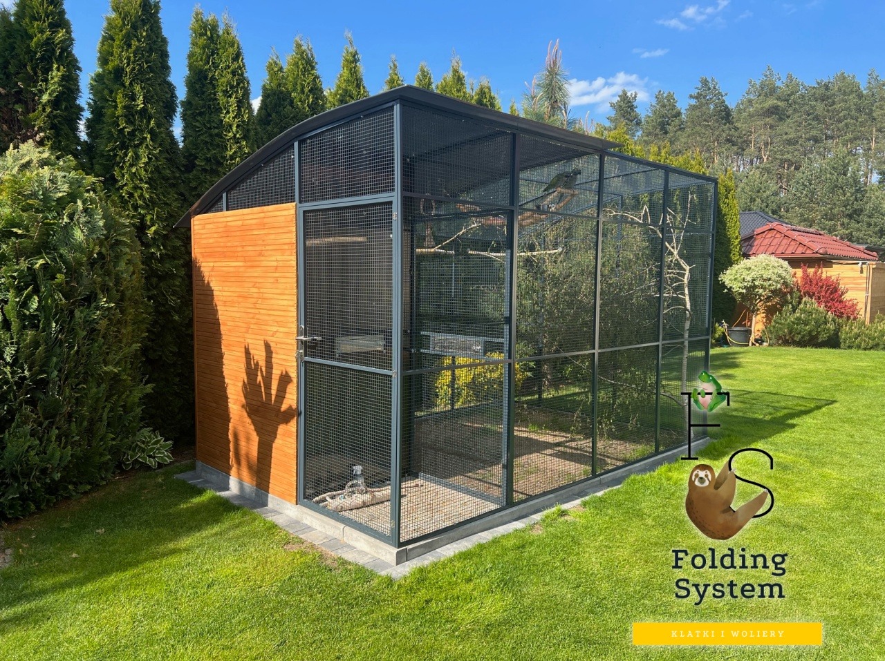 Zewnętrzna, duża woliera z drewnianym panelem i metalową siatką, na trawniku w ogrodzie, z drzewami w tle, oświetlona słońcem.