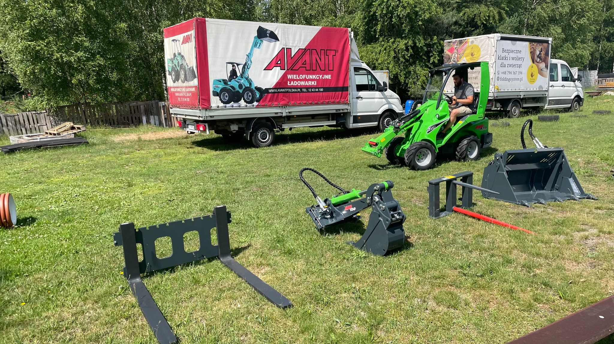 Zielona miniładowarka Avant z operatorem na trawie, z różnymi osprzętami. W tle ciężarówka Avant z reklamą i druga ciężarówka. Słoneczny dzień.