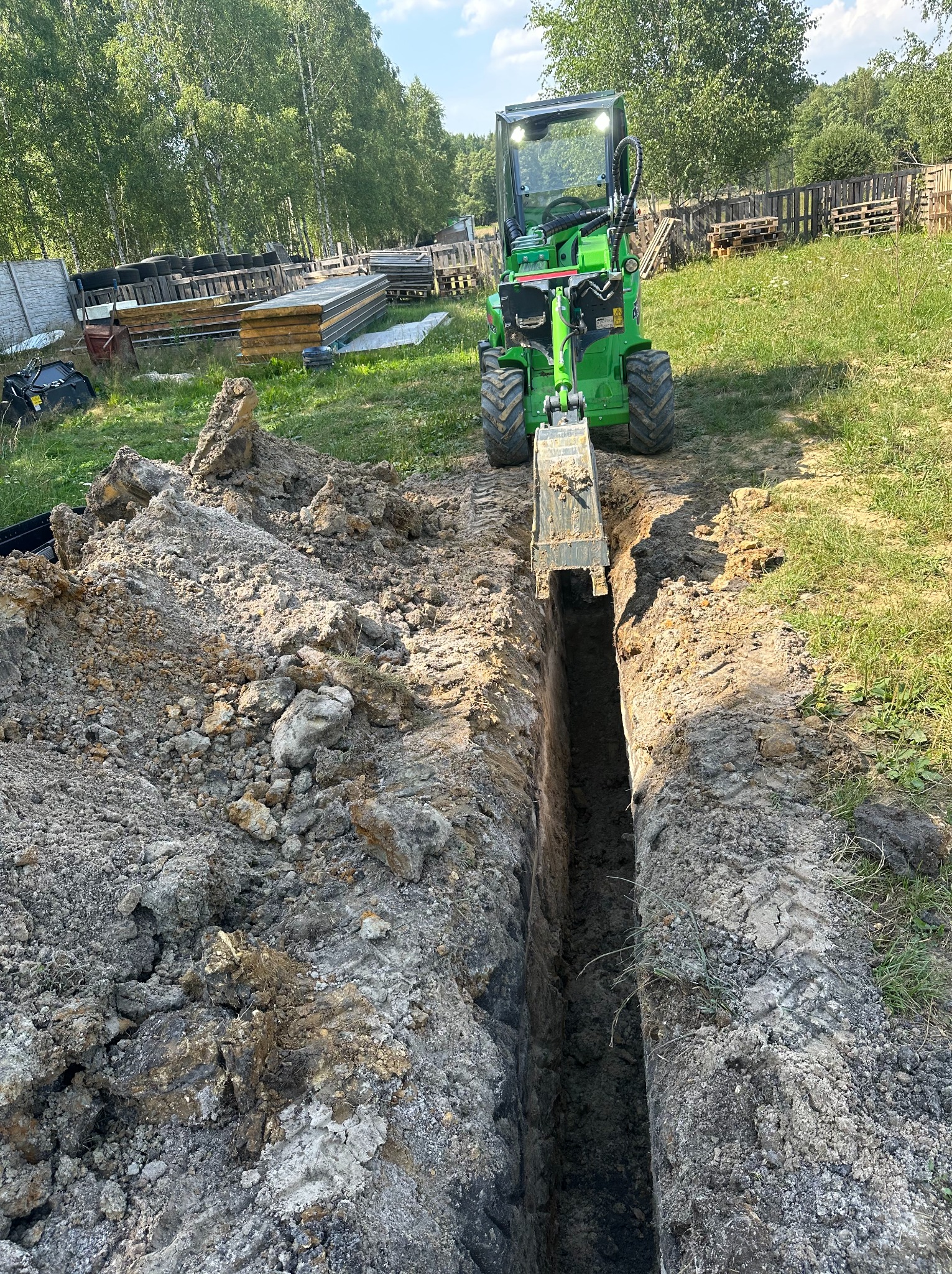 Zielona miniładowarka Avant kopie wąski rów w ziemi w Ossach. Maszyna stoi na trawie, a w tle widać drzewa i materiały budowlane.