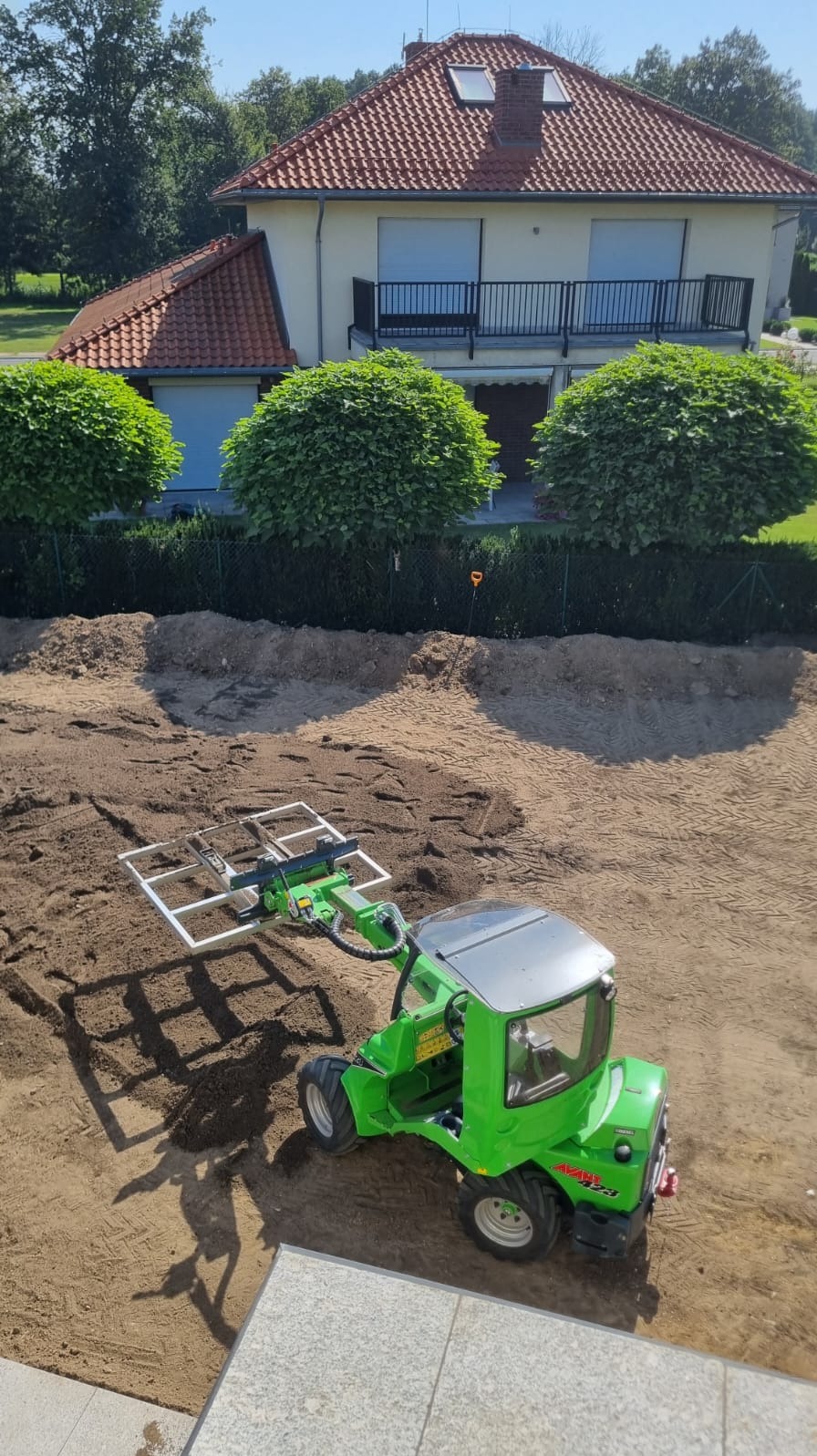Zielona minikoparka Avant z osprzętem do układania kostki brukowej na przygotowanym terenie, w tle dom z czerwonym dachem i kuliste krzewy.