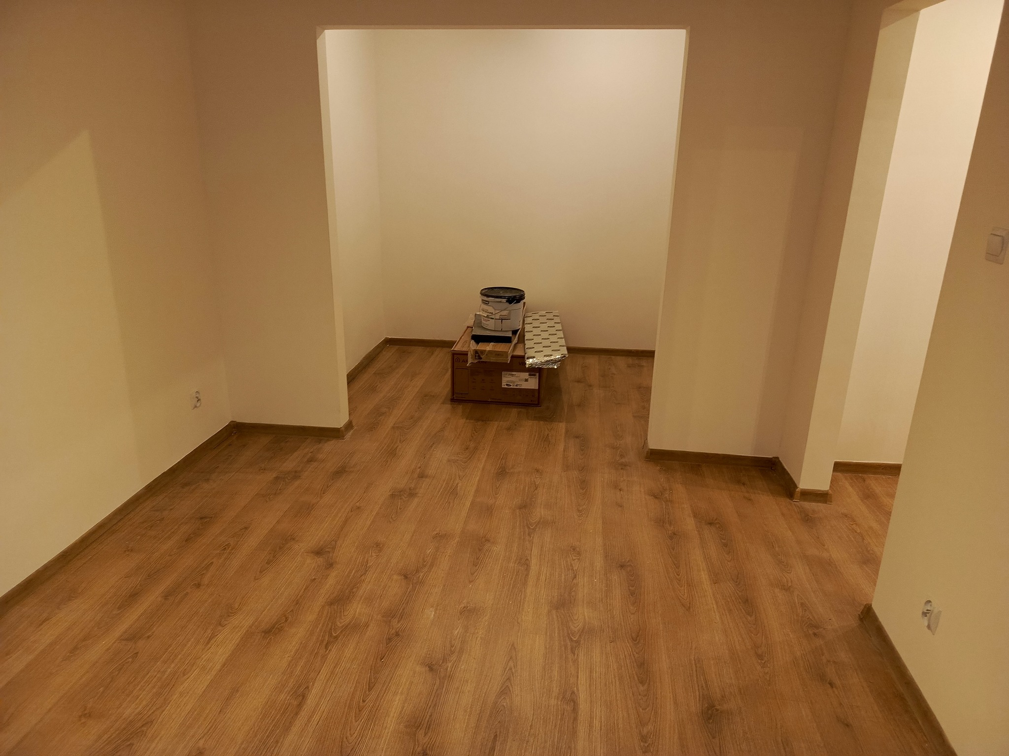 Puste, pomalowane na kremowo wnętrze z drewnianą podłogą. W głębi widoczne puszki z farbą na kartonie, przygotowane do dalszych prac wykończeniowych.
