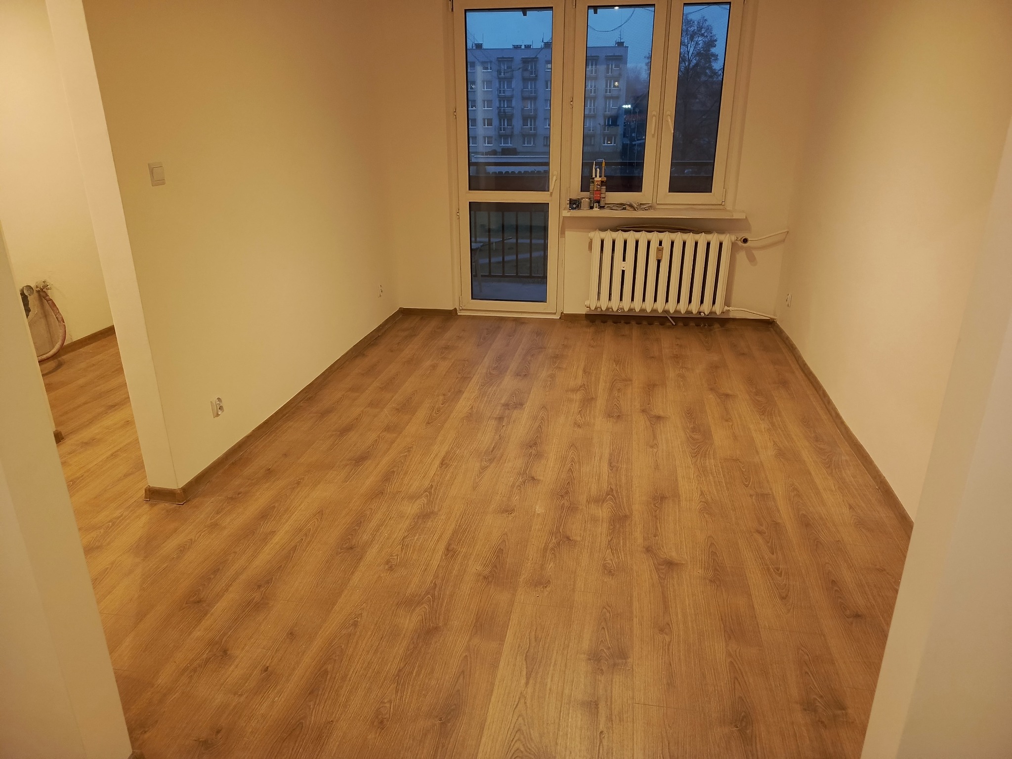 Jasne panele podłogowe w pustym pokoju z białymi ścianami i grzejnikiem pod oknem. Widok na blok zza okna. Świeżo wykończone wnętrze po remoncie.