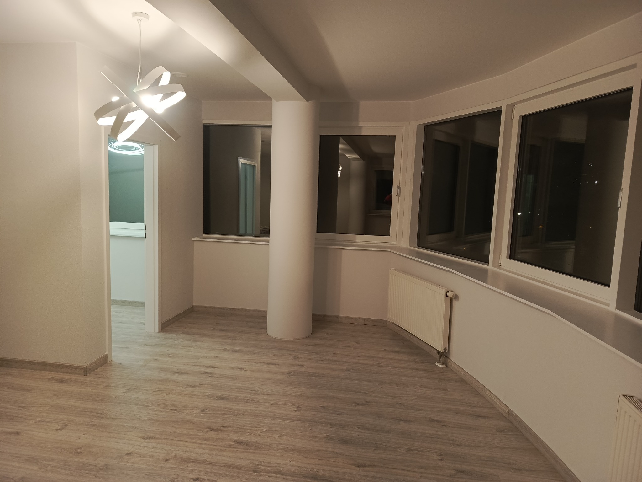 Jasne wnętrze po remoncie: świeżo pomalowane ściany, nowe panele podłogowe, nowoczesna lampa i okna z widokiem na nocne miasto. Minimalistyczny design.