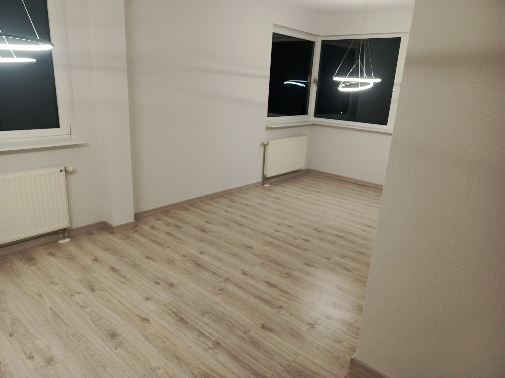 Jasne panele podłogowe w pustym pokoju z białymi ścianami i grzejnikami. Nowoczesne oświetlenie sufitowe. Widok na okno z nocnym krajobrazem.