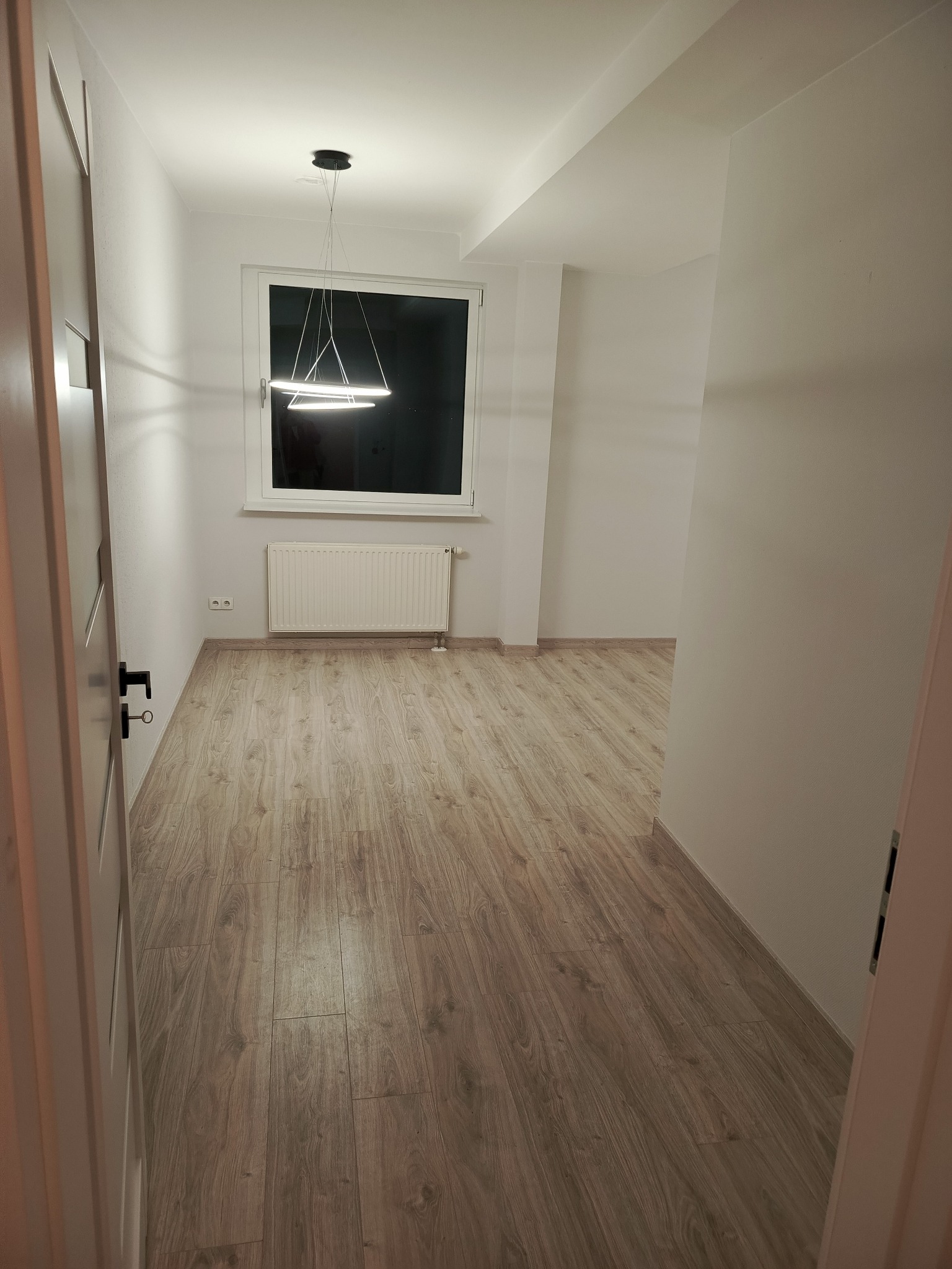 Jasne wnętrze po remoncie: świeżo ułożone panele podłogowe w odcieniach drewna, białe ściany i nowoczesna lampa wisząca. Widok zza uchylonych drzwi.