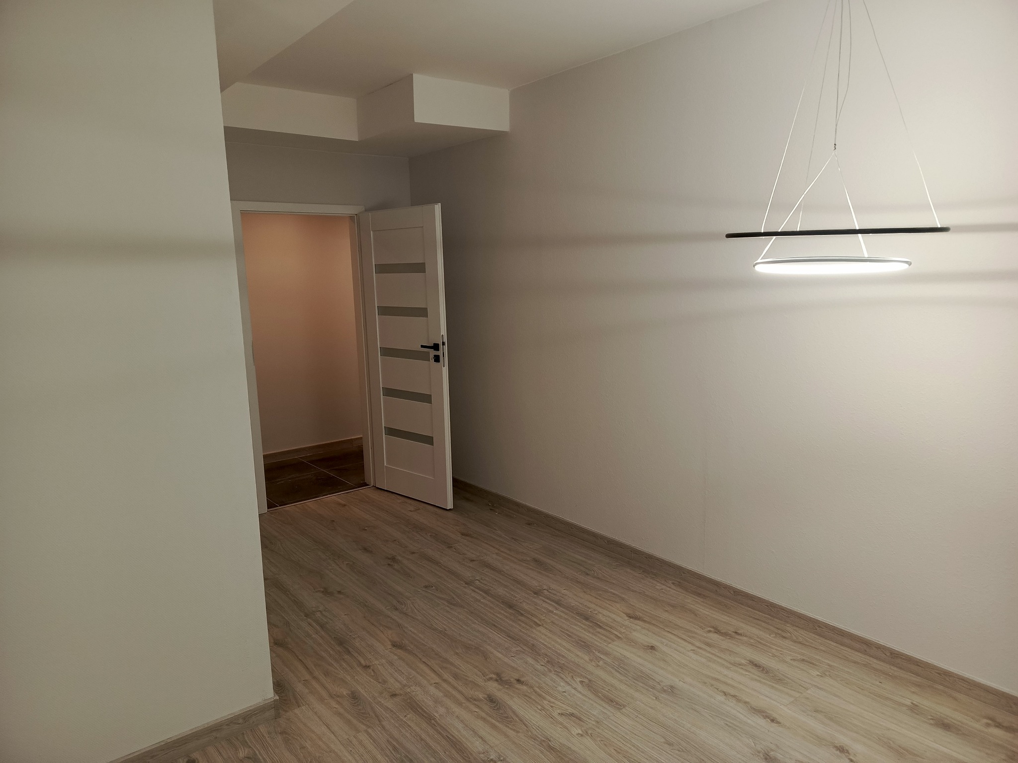 Jasne wnętrze po remoncie: panele podłogowe, pomalowane ściany w odcieniach bieli, nowoczesna lampa wisząca i otwarte drzwi do innego pomieszczenia z ciemną podłogą.