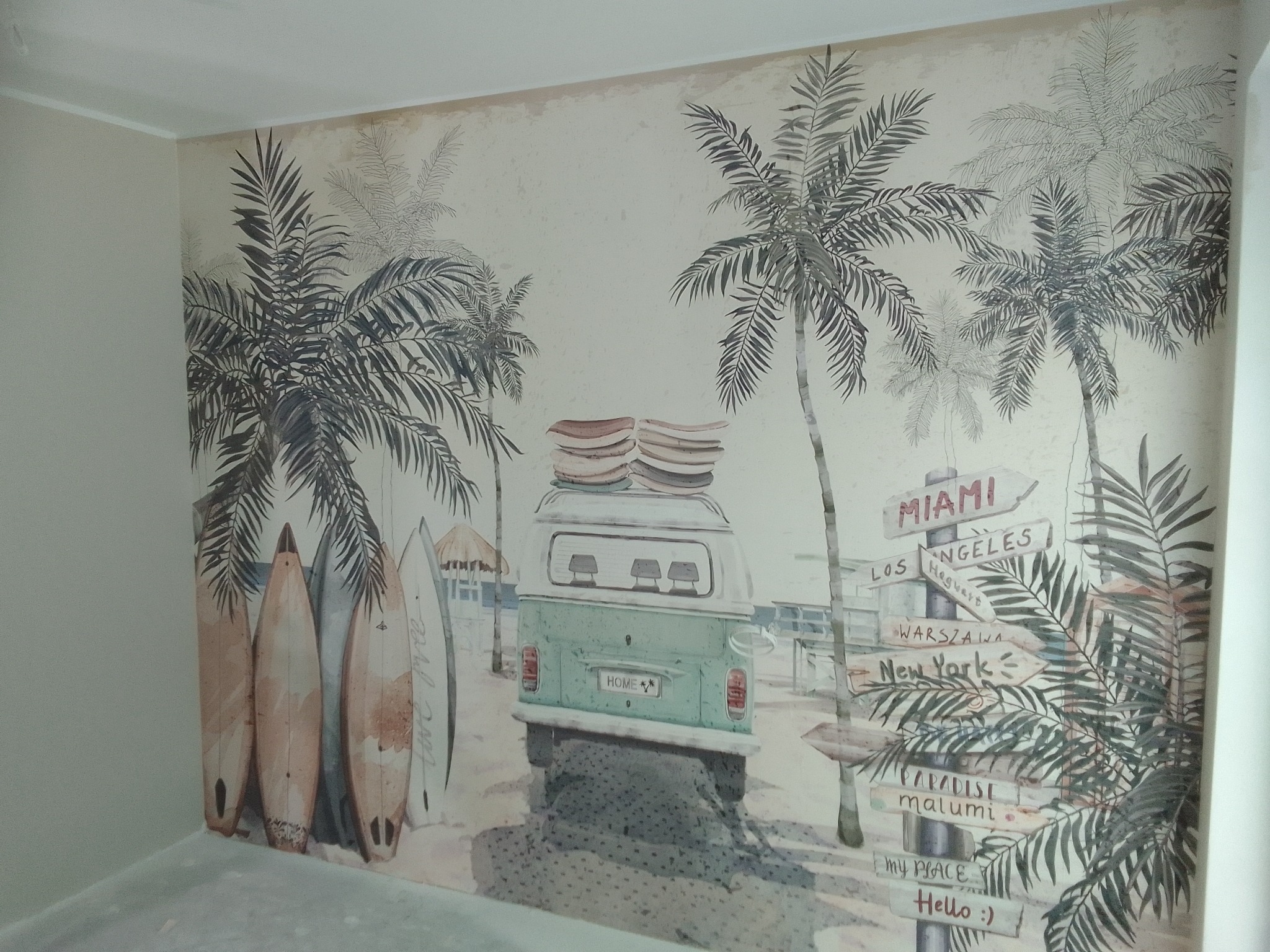 Ściana z fototapetą w stylu vintage, przedstawiającą plażę z palmami, deskami surfingowymi i busem. Dominują stonowane kolory i motywy podróżnicze.