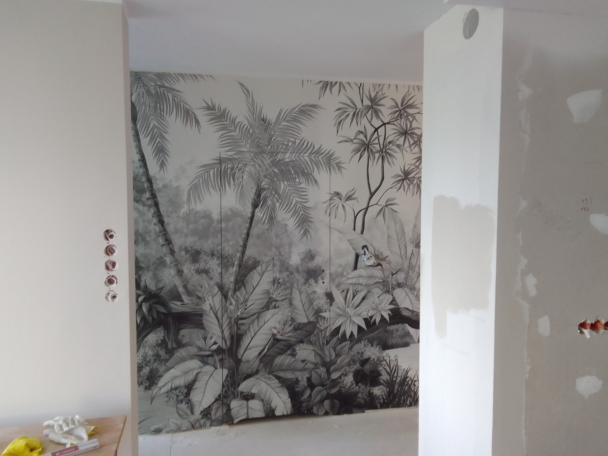 Ściana z fototapetą w motywy tropikalne, widoczne gniazdka elektryczne i fragmenty ścian w trakcie prac wykończeniowych. Realizacja z dbałością o detale.