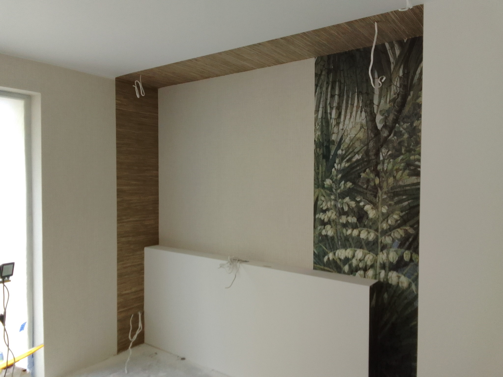 Fragment pokoju po remoncie: ściana z tapetą w motywy roślinne, obok ściana z okładziną imitującą drewno, widoczne kable, surowy stan wykończenia.