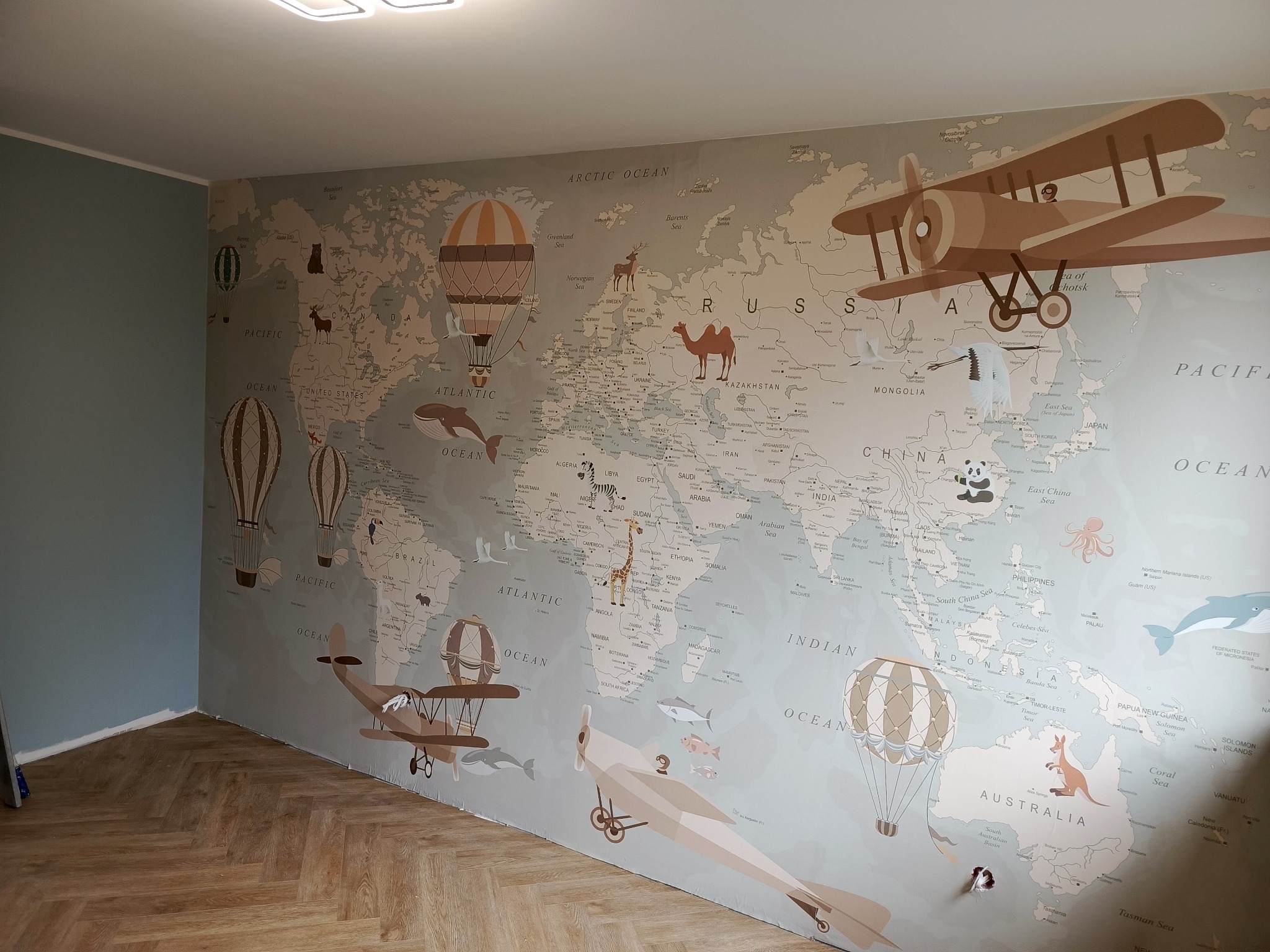 Tapeta z mapą świata w pokoju dziecięcym, z motywami balonów i samolotów, na ścianie pomalowanej na szaro, z parkietem ułożonym w jodełkę na podłodze.