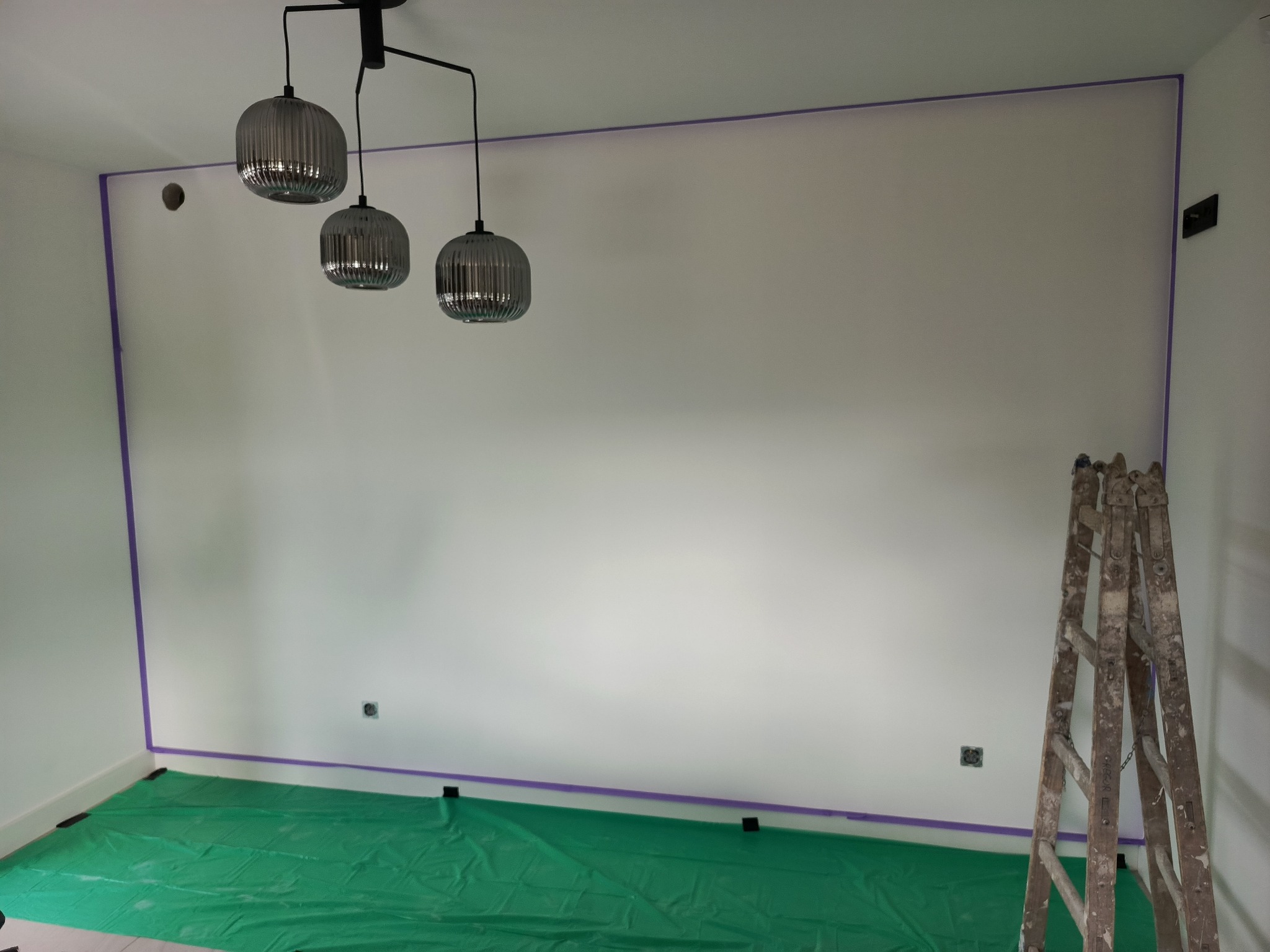 Biała ściana zabezpieczona taśmą malarską, podłoga przykryta folią ochronną, drabina oparta o ścianę. Przygotowanie do malowania.