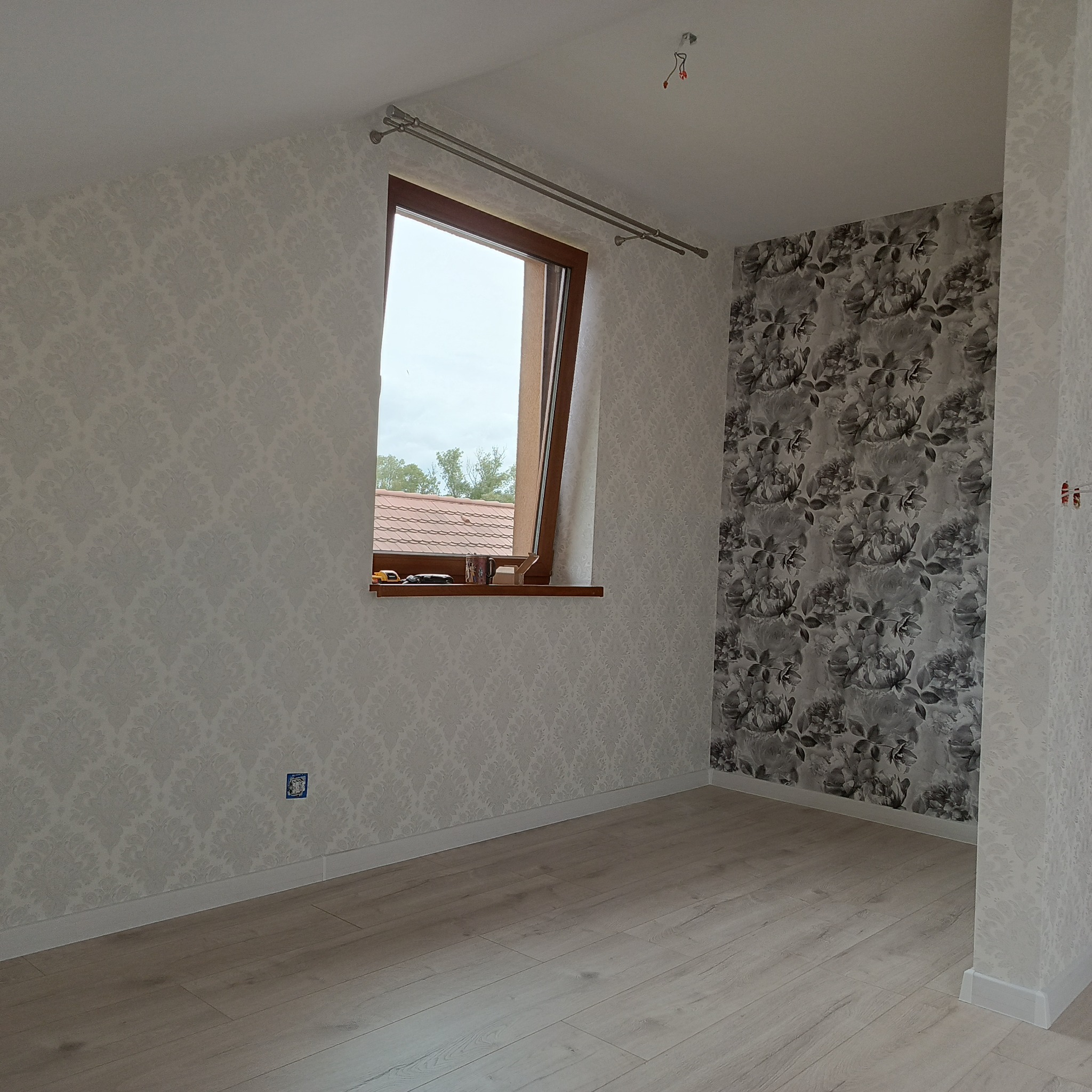 Pokój na poddaszu po remoncie: jasne panele, ściany w dwie różne tapety - wzorzysta szara i biała w ornamenty. Okno dachowe z widokiem na dach.
