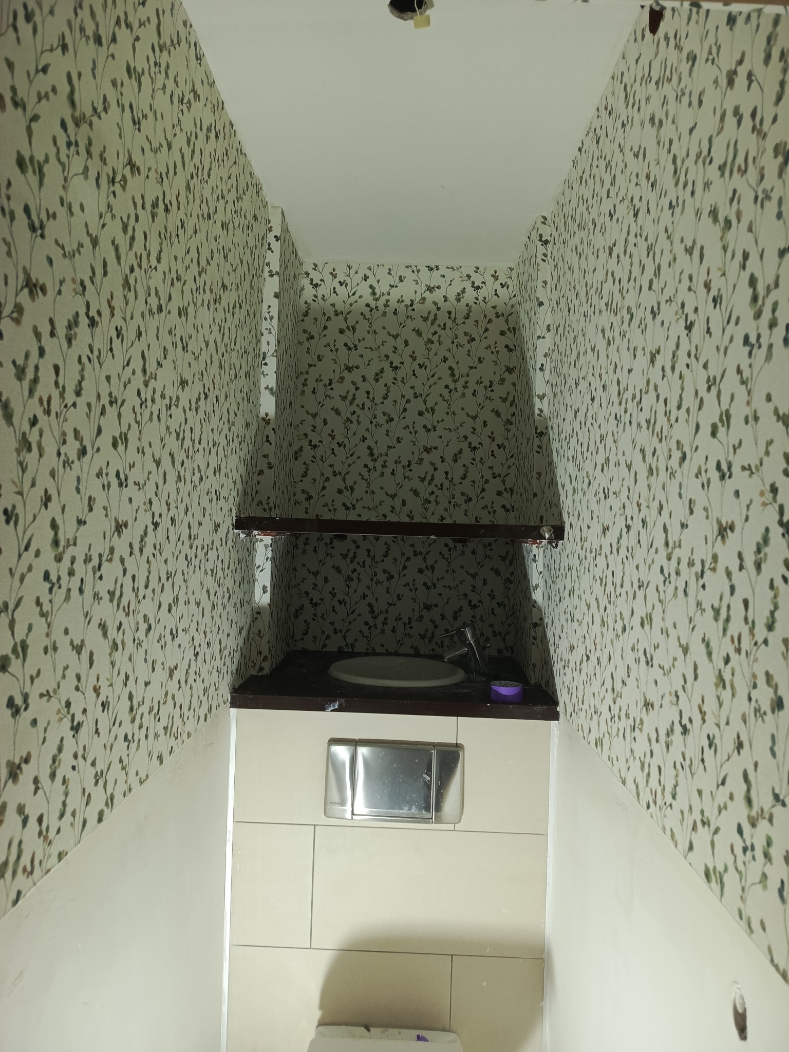 Wykończona toaleta z tapetą w drobne listki, zabudową WC i umywalką. Widok z dołu, ukazujący detale wykończenia i wzór tapety.