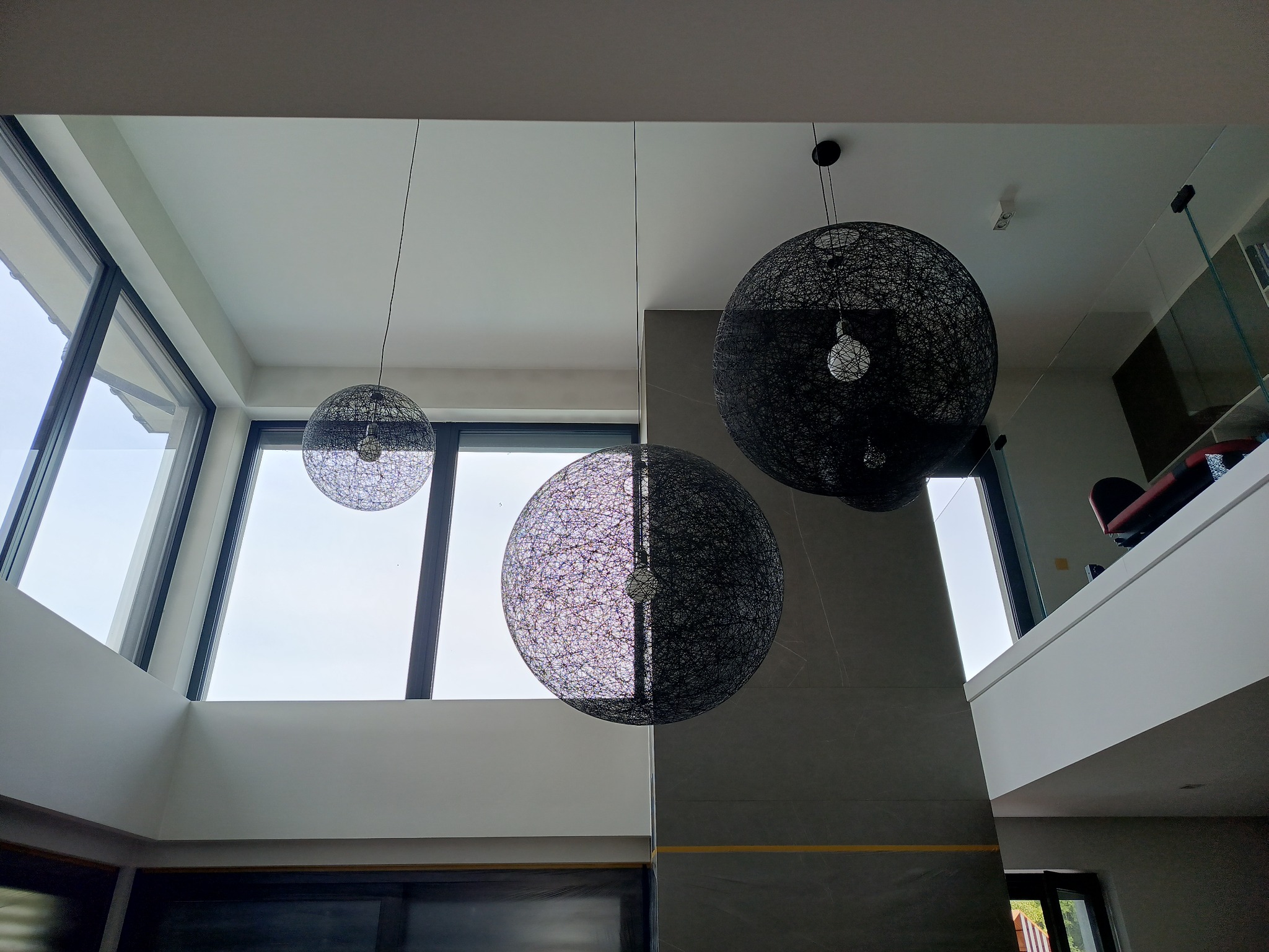 Nowoczesne wnętrze z wysokim sufitem i dużymi oknami, oświetlone trzema kulistymi lampami wiszącymi z czarnego sznurka. Minimalistyczny design, jasne ściany i szare elementy wykończeniowe.