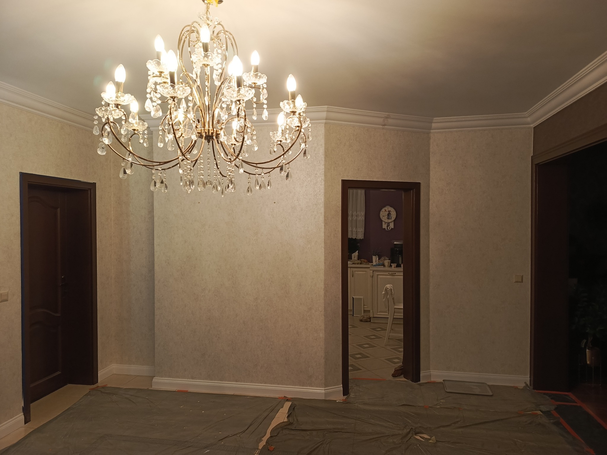 Pokój przed remontem: kryształowy żyrandol, zabezpieczona podłoga, tapeta w delikatny wzór, widoczne listwy przypodłogowe i sztukateria sufitowa. Dwa wejścia.