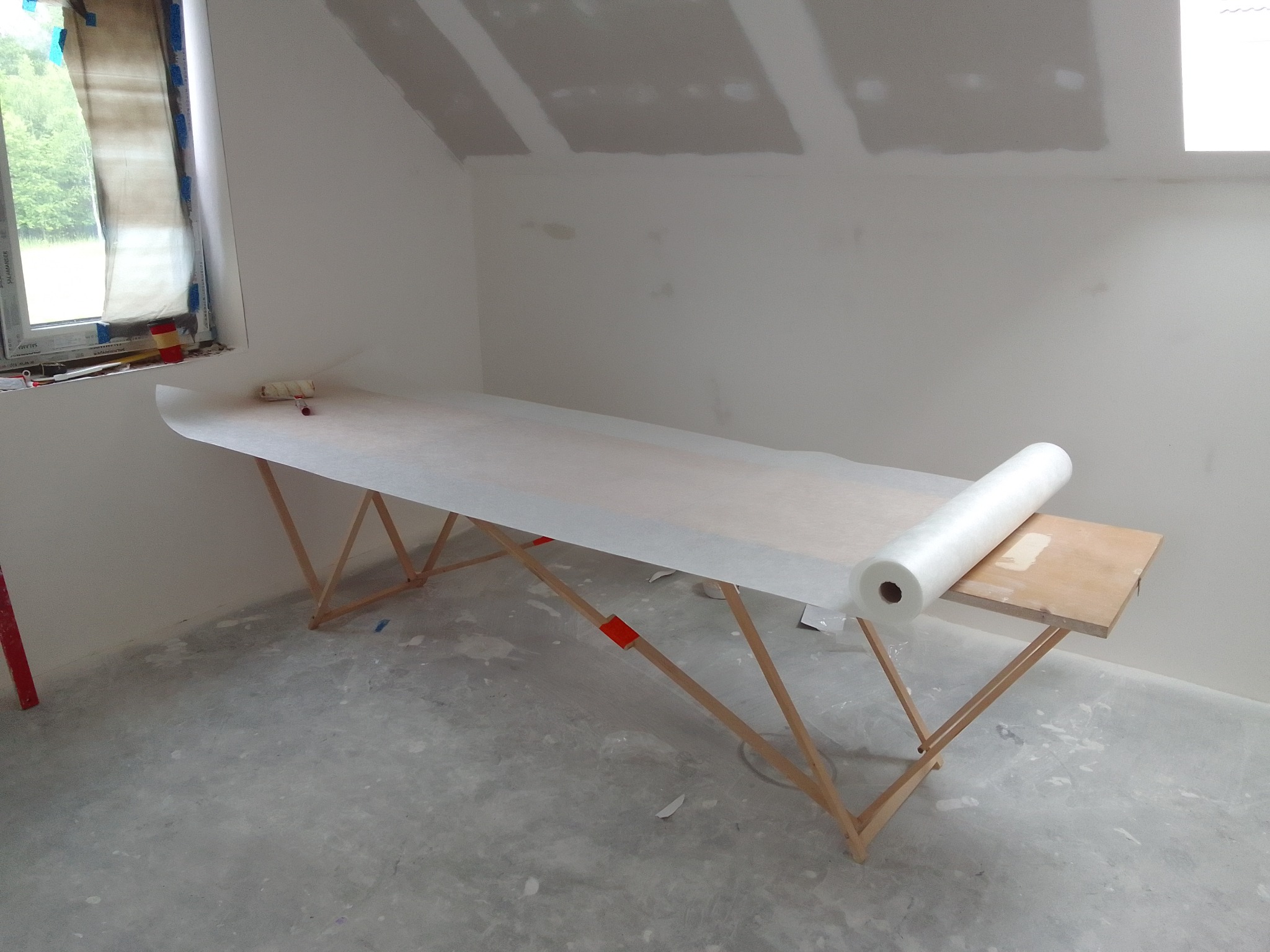 Przygotowanie do tapetowania: stół z rolką tapety flizelinowej w surowym wnętrzu z oknem i płytami gips-karton na poddaszu. Widoczne narzędzia i niedokończona podłoga.