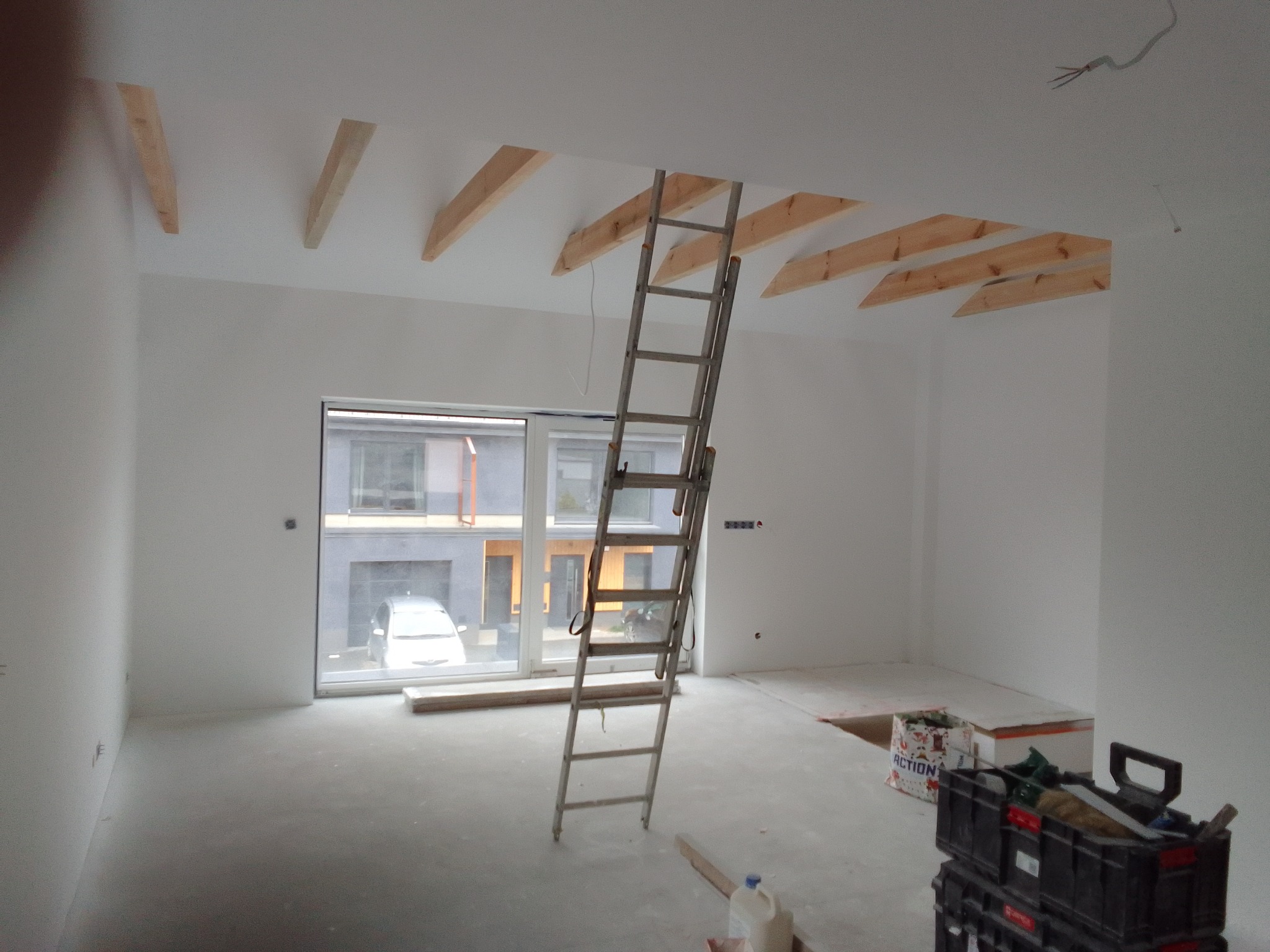 Wnętrze w trakcie prac wykończeniowych: surowe drewniane elementy na suficie, drabina oparta o ścianę, narzędzia i materiały budowlane. Widok na okno i otoczenie.