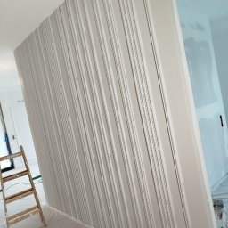 Adrianzlotaraczka - Ściana z dekoracyjnymi listwami w odcieniach szarości, częściowo pomalowana. Widoczna drabina malarska i zabezpieczona folią podłoga. Prace wykończeniowe w trakcie.