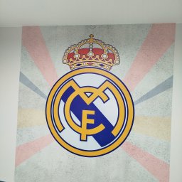 Adrianzlotaraczka - Malowidło na ścianie z herbem Realu Madryt i koroną. Tło w paski w odcieniach beżu i szarości. Fragment niebieskiej kanapy w dolnej części kadru.