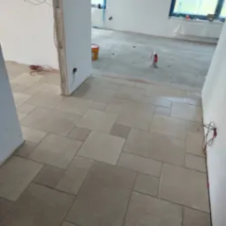 Ukończona podłoga z beżowych płytek ułożonych w geometryczny wzór. Widoczne białe ściany w trakcie wykończenia i okno z niebieską folią ochronną.