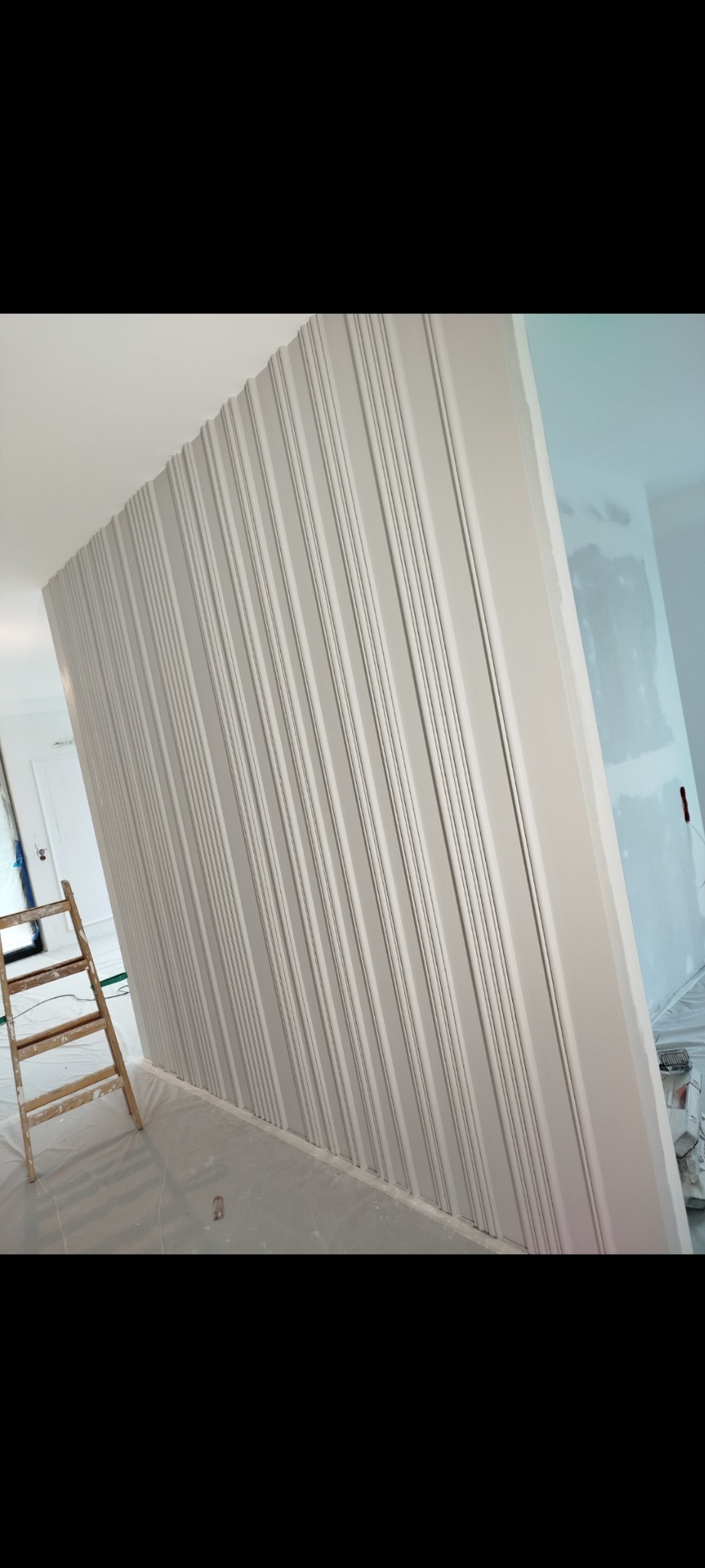 Ściana z dekoracyjnymi listwami w odcieniach szarości, częściowo pomalowana. Widoczna drabina malarska i zabezpieczona folią podłoga. Prace wykończeniowe w trakcie.