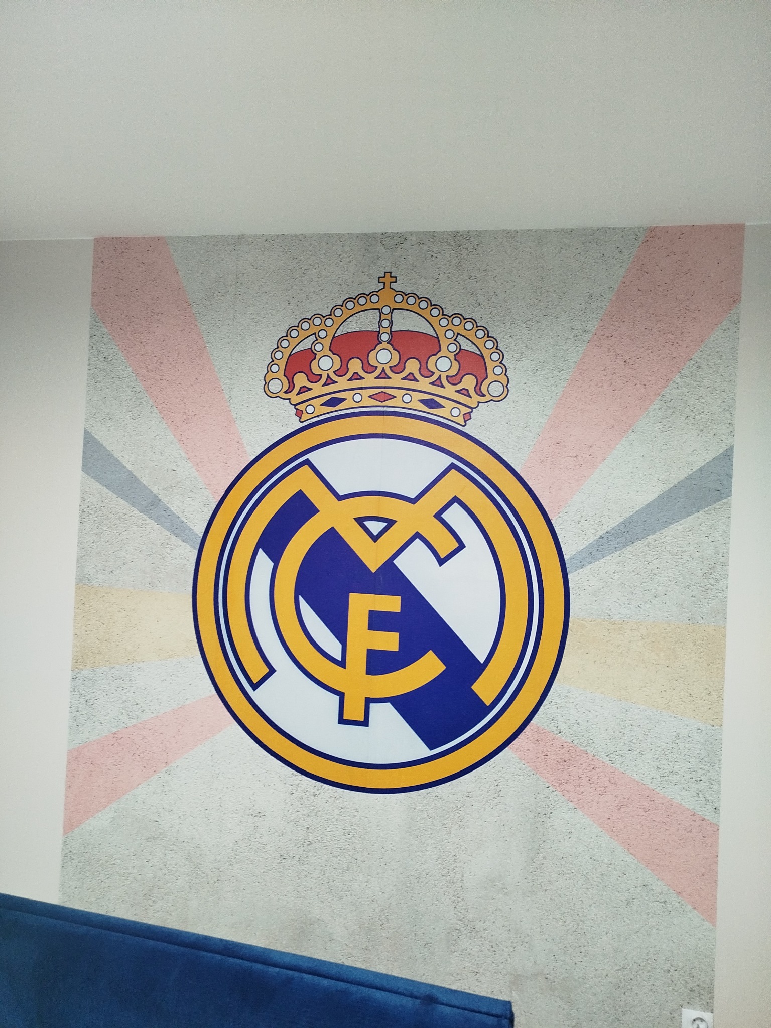 Malowidło na ścianie z herbem Realu Madryt i koroną. Tło w paski w odcieniach beżu i szarości. Fragment niebieskiej kanapy w dolnej części kadru.