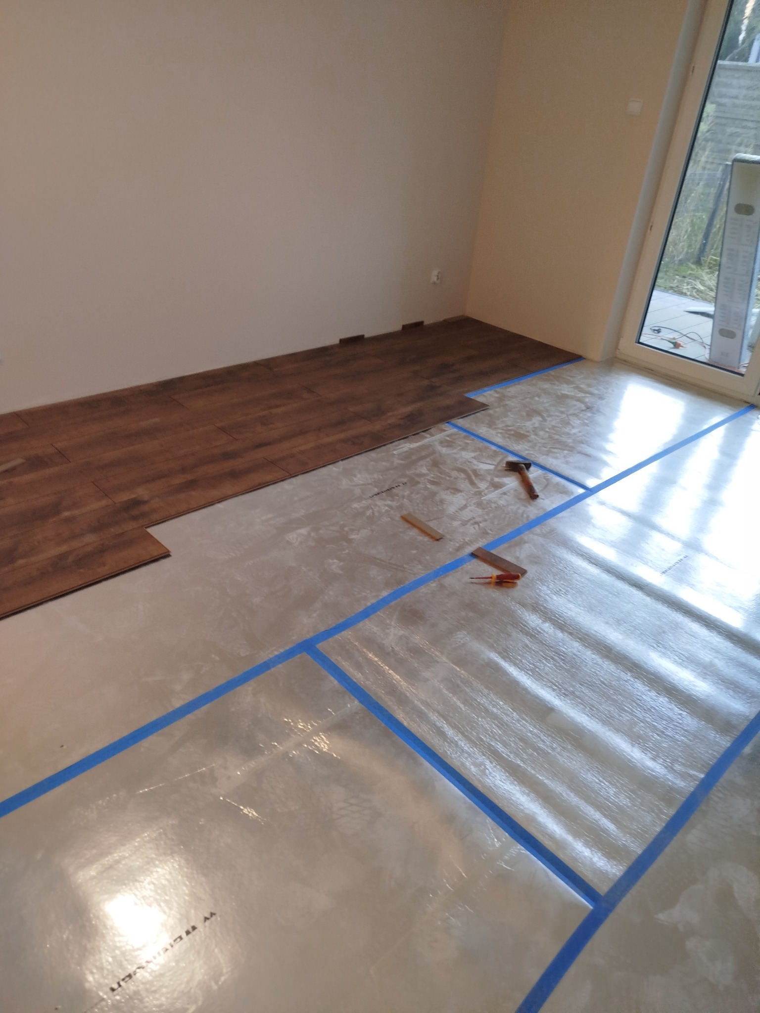 Układanie paneli podłogowych w pokoju, częściowo zakończone. Widoczne narzędzia: młotek, dłuto i ołówek. Podłoże zabezpieczone folią i taśmą. Ujęcie z góry.