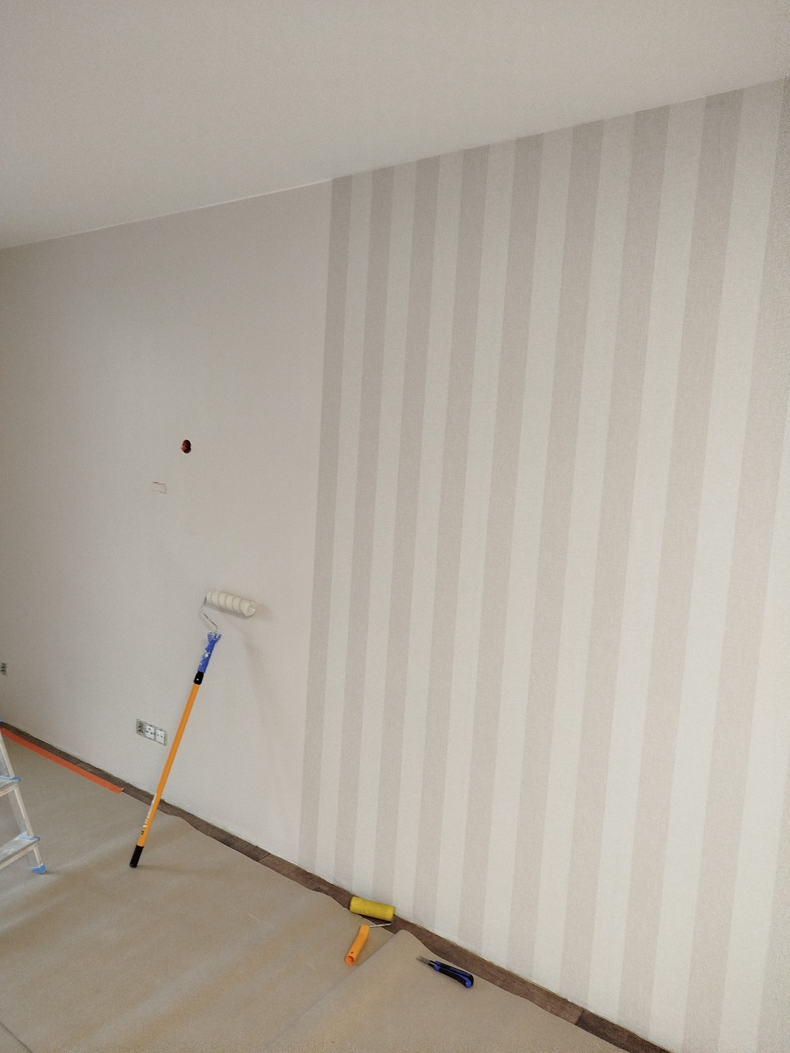 Ściana w trakcie malowania w pionowe pasy jasnymi odcieniami, wałek oparty o ścianę, podłoga zabezpieczona folią. Widoczne narzędzia malarskie.