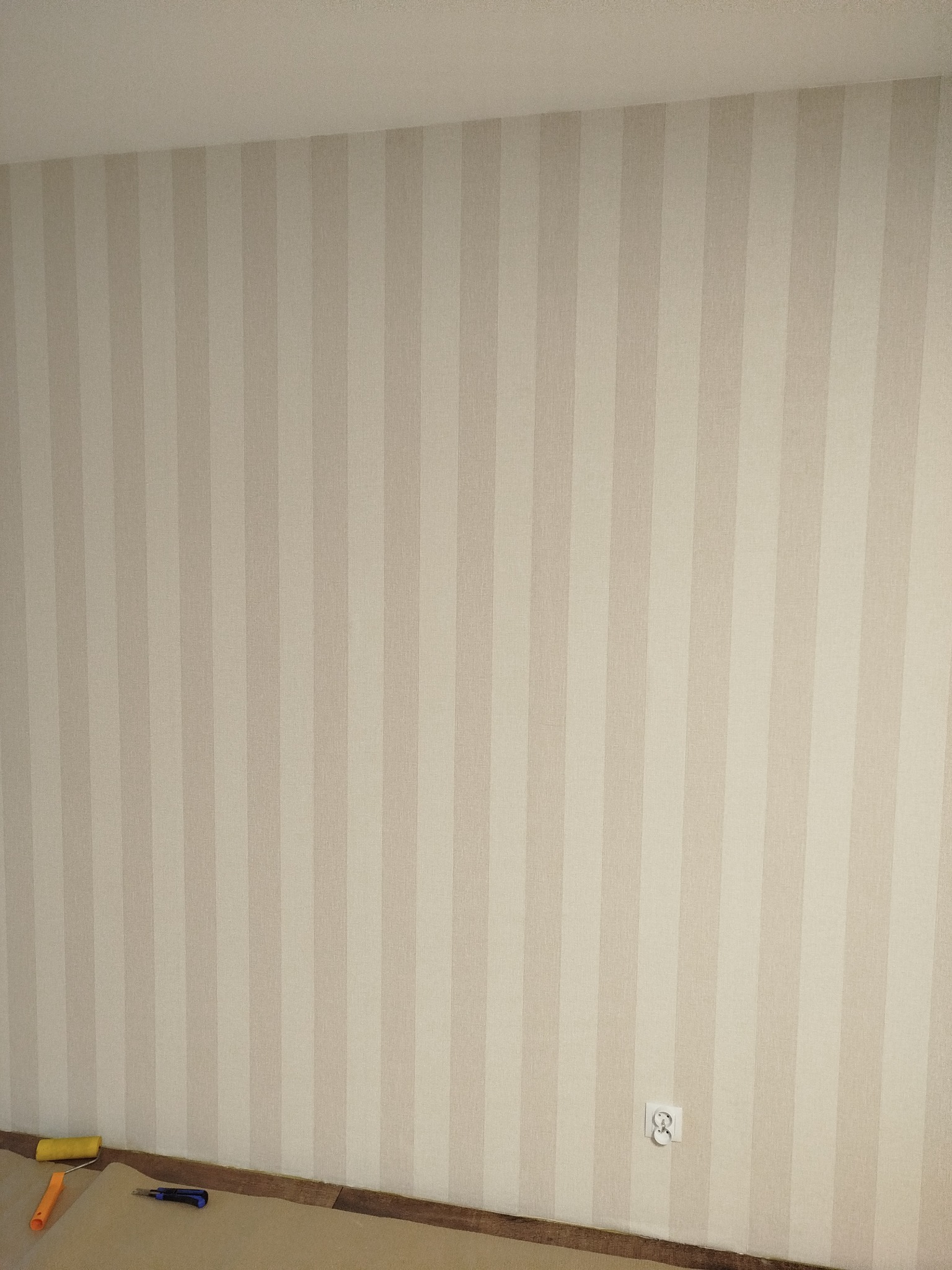 Ściana w beżowe pasy po tapetowaniu; widoczny fragment podłogi z narzędziami: wałek i nóż. Gniazdko elektryczne na ścianie. Stonowana kolorystyka.