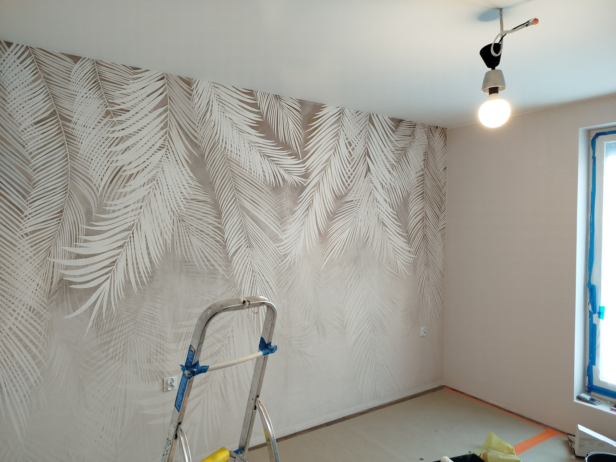 Pokój po tapetowaniu w liście palmowe, z drabiną i prowizorycznym oświetleniem sufitowym. Widoczne przygotowania do dalszych prac wykończeniowych.