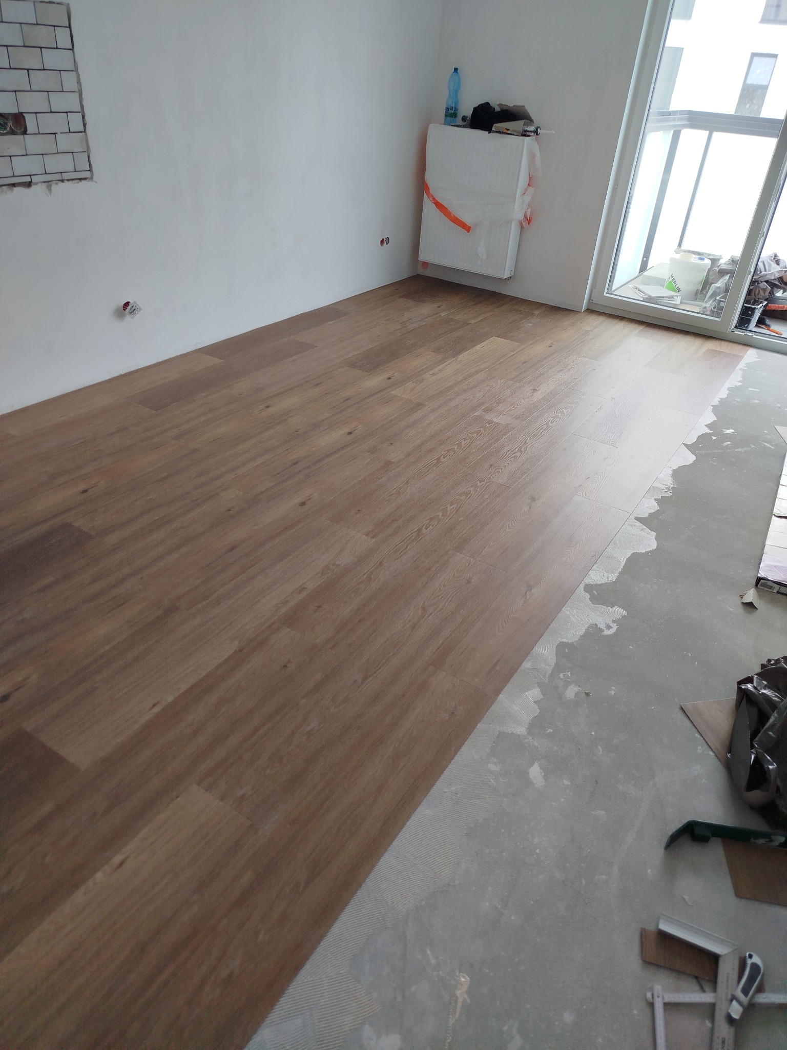 Ułożone panele podłogowe w nowym wnętrzu, widoczne narzędzia i resztki materiałów budowlanych. Częściowo wykończone ściany i duże okno.